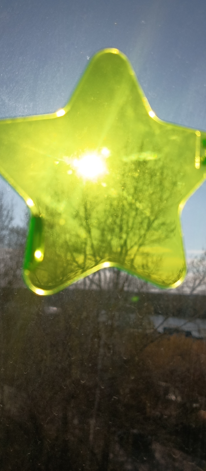 The image shows a green star-shaped object affixed to a window. Sunlight shines through the star, creating a bright glare. A blurry view of trees without leaves is visible through the star and outside the window. Buildings are faintly visible in the background beyond the trees. The location is unidentified.