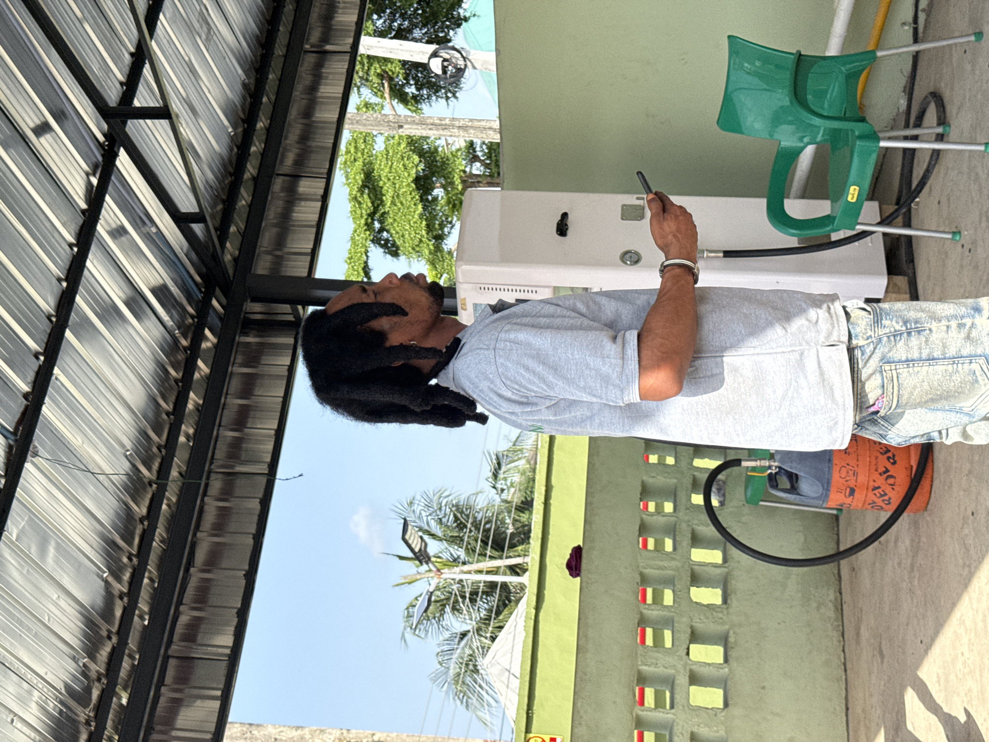 A man with dreadlocks is standing next to a white appliance and a green child's chair, outdoors. He is wearing a light grey shirt and blue jeans. He is holding a black cell phone in his right hand. The ground is concrete. Behind the man is a green wall with decorative rectangular cutouts. In the background, there are trees and a blue sky with some clouds. A metal awning is partially visible in the top left corner of the frame. A 10 kg orange cooking gas cylinder and hose are connected to the appliance next to him. Location is likely in Ado, Nigeria, though the exact area is not discernible.