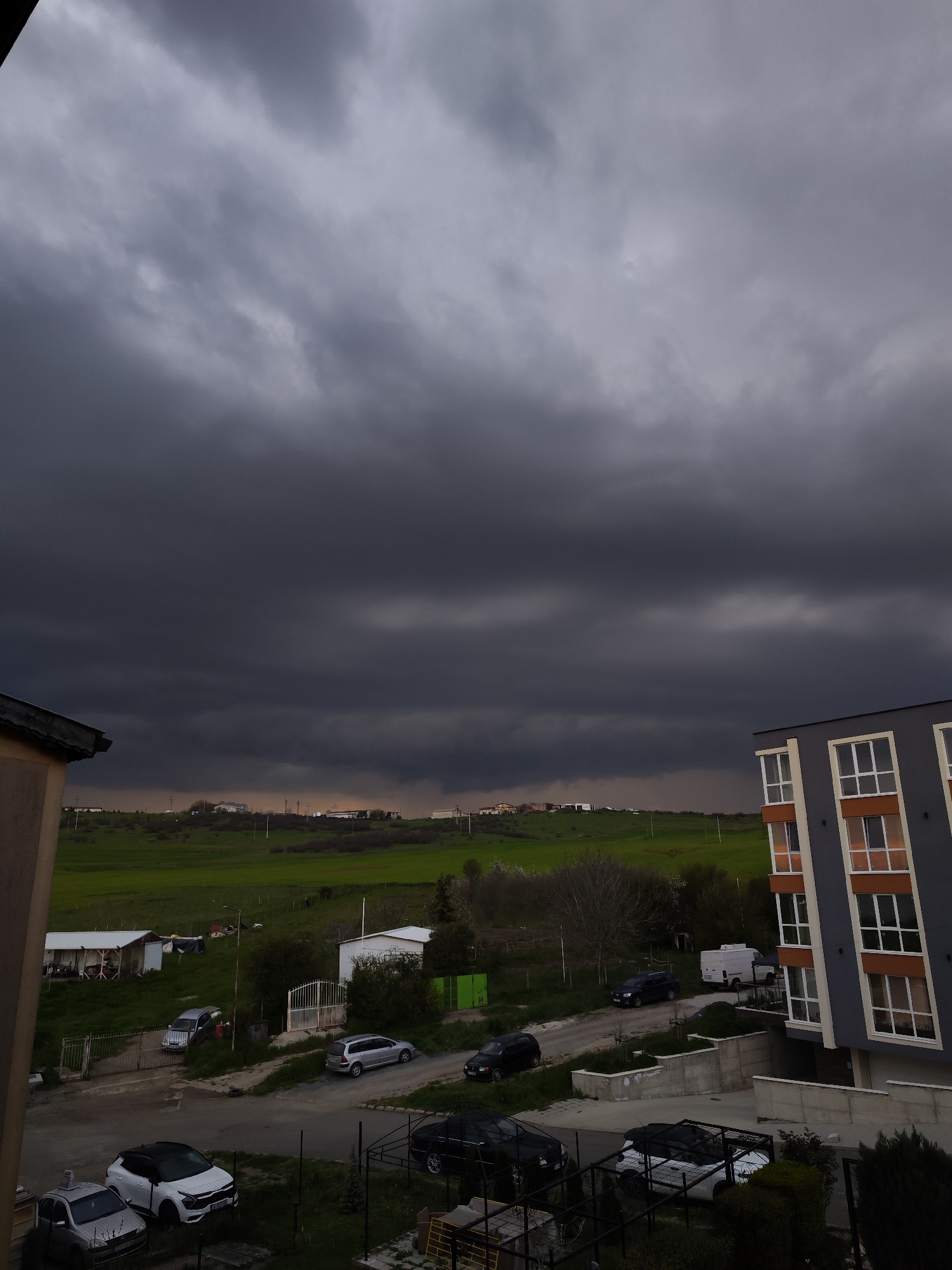 The image captures a somber, overcast landscape from an elevated perspective, likely a balcony or upper floor of a building. The dominant feature is the sky, filled with dense, dark grey to almost black storm clouds that stretch across the entire upper half of the frame, indicating impending rain or a heavy storm. A faint band of lighter, possibly yellowish light is visible very low on the horizon in the distance, contrasting with the dark sky.
The scene depicts a residential area bordering a green, hilly countryside. In the foreground, parts of two buildings are visible: a light brown structure on the left and a modern, multi-story apartment building with dark grey/blue walls and distinctive white-framed windows with orange-brown accents on the right.
The midground features a mixture of green grass and dirt tracks, with several parked cars, including a white compact SUV (possibly a Kia Seltos/Celtos) with a partially visible Bulgarian license plate "CA 6...", a silver hatchback, a dark sedan, a silver station wagon, and a white delivery van. Scattered amidst the greenery are small, functional-looking structures, possibly sheds or workshops, and various trees and bushes. Fences and gates delineate properties.
In the background, rolling green hills rise, dotted with a few distant buildings and utility poles and lines. The overall impression is one of low light, characteristic of a heavily cloudy day or twilight just before a storm, creating a moody and atmospheric setting in what appears to be an outskirts or semi-rural area near Burgas, Bulgaria. No people are visible, and no specific activity is taking place.