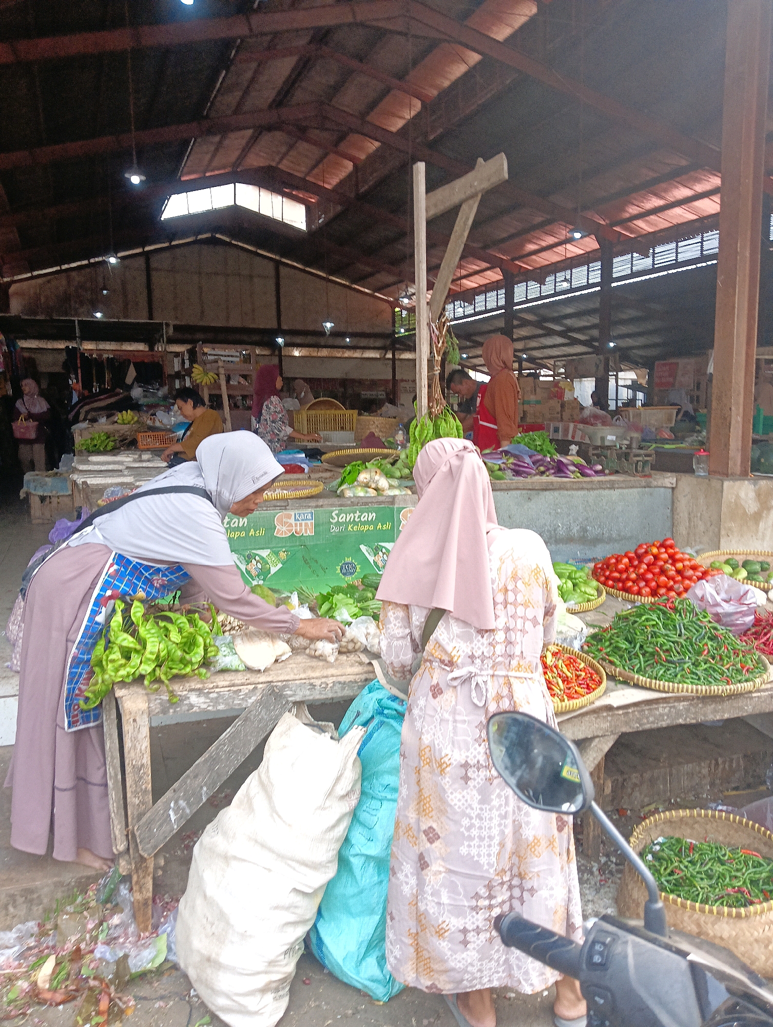 This is a full-shot, eye-level, natural light image of a bustling outdoor market in Sokaraja, Indonesia. The scene is dominated by stalls laden with fresh produce. In the foreground, two women in hijabs are seen interacting at a market stall. The woman on the left, wearing a light grey hijab and a brown and blue patterned apron over a purple abaya, is reaching towards a pile of green beans. The woman on the right, wearing a light pink hijab and a patterned dress, is also looking at the produce. A motorcycle is parked to the right of the frame, its side mirror reflecting the scene.
Behind the women, various vegetables are displayed, including tomatoes, green and red chilies, eggplants, and leafy greens. Large woven baskets and plastic bags are used to hold and display the produce. Bags of what appear to be produce or other market goods are stacked on the ground. The market appears to be covered by a large, weathered roof structure supported by metal beams, with open sides allowing natural light to filter in. Other market-goers are visible in the background, as well as more stalls with produce and goods. The ground is concrete and appears somewhat worn and dirty, with discarded vegetable scraps and plastic debris scattered around. The overall atmosphere is one of active commerce and daily life in a local Indonesian market.
