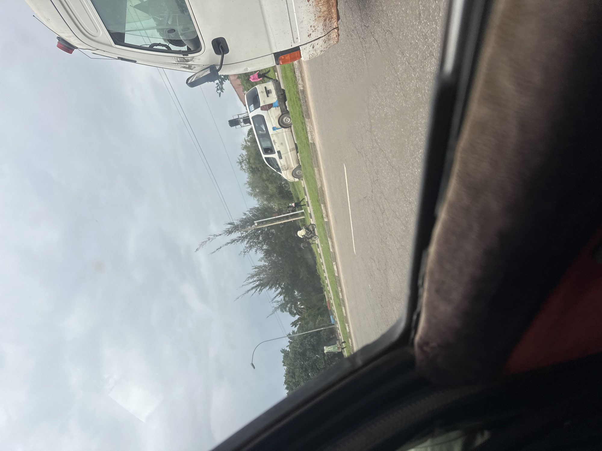 The image shows a view from inside a vehicle, looking out at a street scene in Aso, Nigeria. The sky is overcast, suggesting a cloudy or potentially rainy day. The scene appears to be during daylight hours, likely morning or afternoon.
Several vehicles are visible, including a white van in the foreground and another white van further down the road. One of the vans has visible dirt and rust on its exterior. In the distance, a motorcycle is being ridden. There are also several people visible. One person in a pink garment is standing near the side of the road. Another person appears to be walking or standing on the side of the road, wearing a green shirt. Trees and vegetation line the side of the road, indicating a relatively green area. A lamppost is also present. The road itself appears to be paved.
The image is oriented vertically, with the top of the frame showing the sky and the bottom showing the interior of the vehicle from which the picture is taken. The perspective suggests the vehicle is in motion or stopped on the side of the road. There is no visible text in the image. The overall impression is a common street scene in a Nigerian city on a cloudy day, with everyday activities and vehicles present.