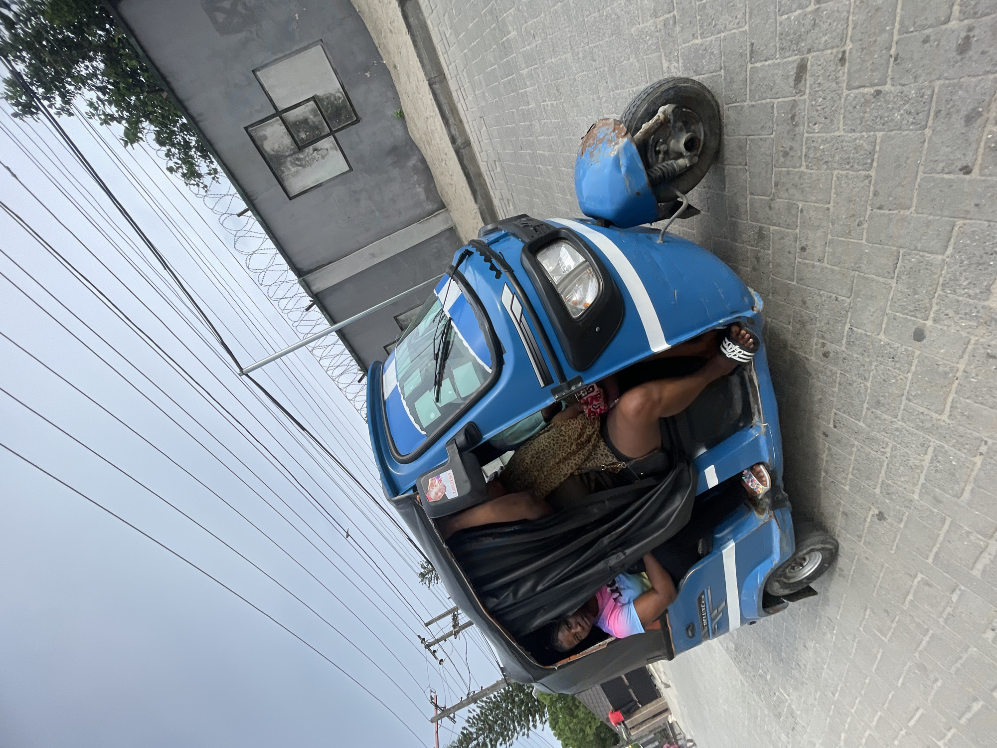 The image, taken in Iranla, Nigeria, shows a blue three-wheeled auto-rickshaw (commonly known as a Keke Napep) parked on a paved street. The vehicle is positioned next to a grey building or wall, which is topped with razor wire.
Inside the auto-rickshaw, two individuals are visible. In the front passenger area, a person is seen wearing a leopard-print top and black and white striped sandals, with their legs extended. Further back, another person, wearing a colorful tie-dye t-shirt, looks directly at the camera with a neutral expression. The interior of the vehicle has black fabric or tarp coverings. A small framed picture, with the text "PROPERTY OF..." partially visible, is mounted inside near the front. The blue vehicle has white stripes and exhibits some signs of wear, including rust on the front fender.
The sky overhead is overcast, indicating a cloudy daytime setting. Numerous utility lines stretch horizontally across the frame. Some green foliage from trees can be seen in the upper left. The paved surface beneath the vehicle resembles grey cobblestones or pavers. The overall scene captures a candid moment of daily life in a Nigerian urban or semi-urban environment.