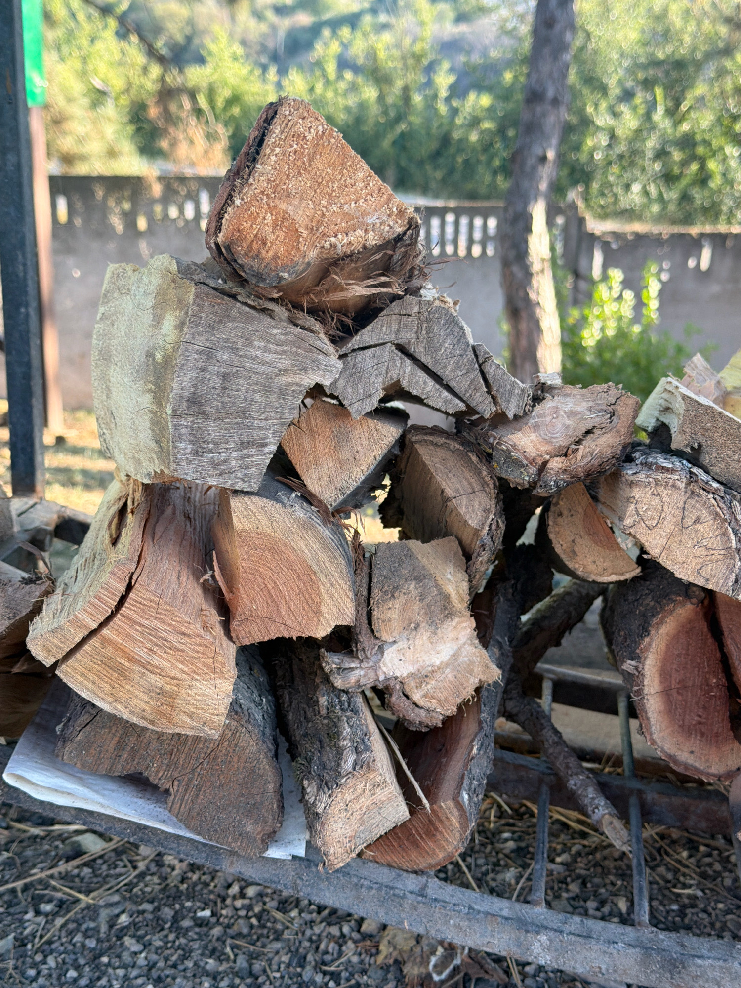 A close-up, eye-level, outdoor shot shows a pile of chopped firewood stacked on a metal grate. The wood varies in size and shape, with some pieces exhibiting rough bark and others showing the smooth, aged grain of cut timber. The firewood is predominantly light brown and gray, with some pieces displaying darker tones and visible cracks. In the background, a blurry green landscape with trees and foliage is visible, along with a low, light-colored wall with vertical openings. Sunlight casts dappled shadows, suggesting it is daytime. The ground beneath the grate is covered in small stones and dry vegetation. There are no people or animals present in the image. The scene is located in the city of Perovske, Ukraine.