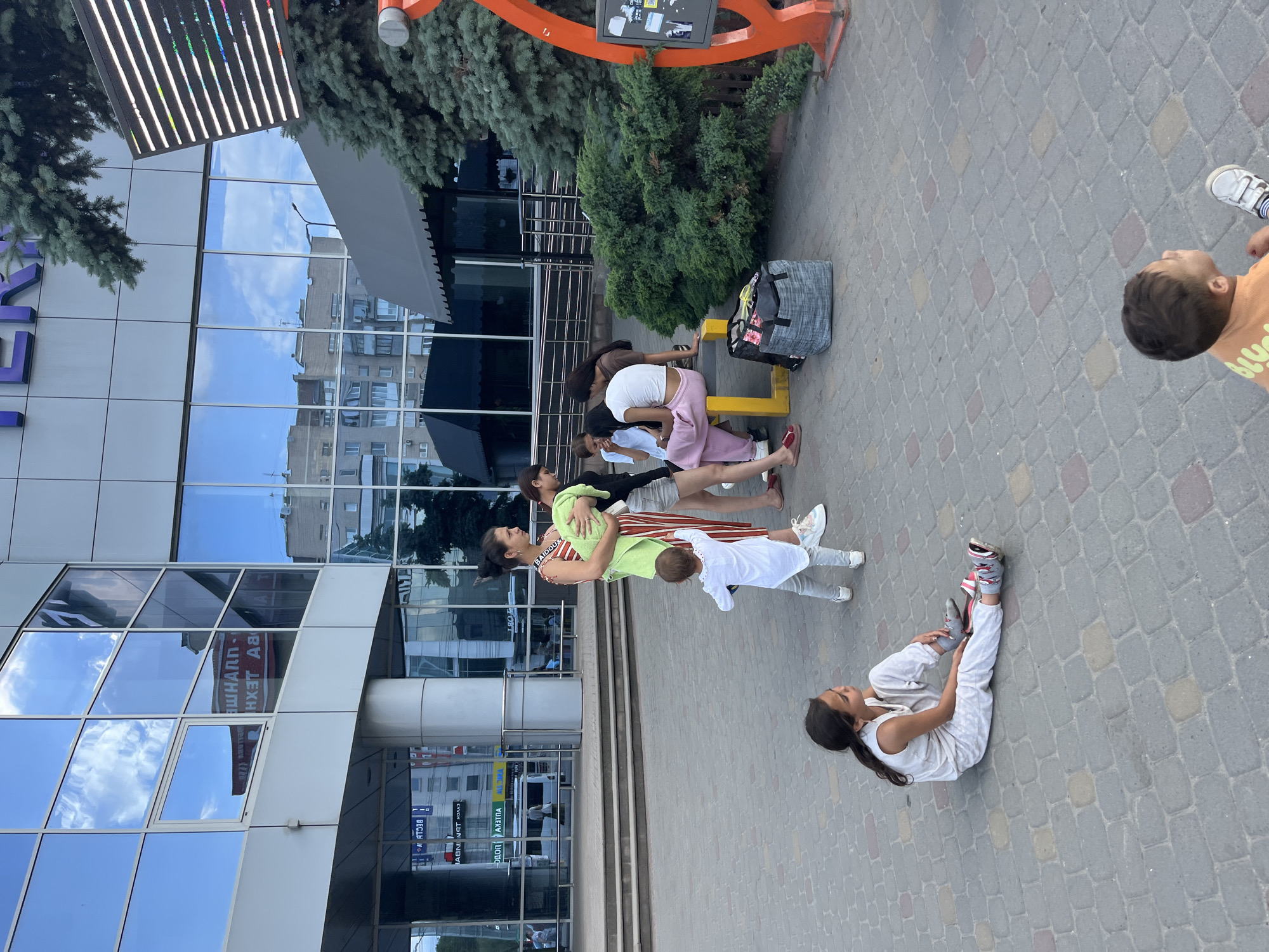 A group of people, including children and adults, are gathered outside a modern building with large glass windows that reflect the sky and surrounding structures. The building has a gray awning extending over the entrance area. The ground is paved with interlocking bricks. Several individuals are in various poses, some sitting or lying down on the pavement, while others are standing or engaged in an activity that appears to be some form of exercise or stretching. One woman in a colorful striped dress and a green wrap is holding a child. Another child in an orange shirt is looking up. A young girl in a white sleeveless top and pants is lying on the ground, stretching her legs. A large grey duffel bag is visible near a yellow structure. The overall atmosphere seems relaxed, and the time of day appears to be daytime, possibly late afternoon, given the lighting. Text is visible on the building's windows, some of which is reversed due to reflection. The text appears to be in Cyrillic script and includes words like "ВАЙБОЦ", "ДУМ", and "АНД".
