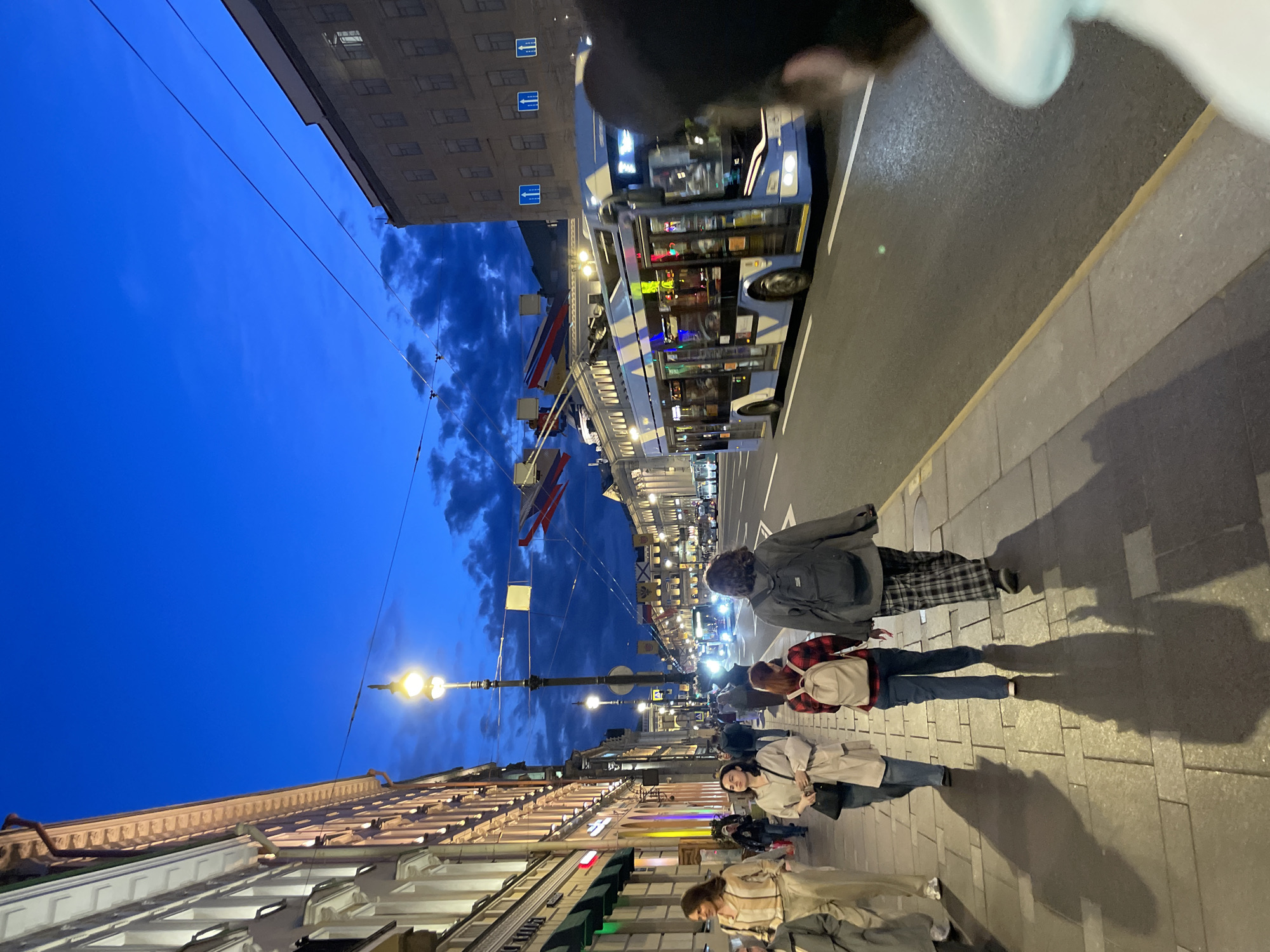 An evening street scene, likely in Saint Petersburg, Russia, captures a bustling thoroughfare under a deep twilight sky. The image, when viewed upright, looks down a wide city street flanked by multi-story buildings and illuminated by streetlights.
In the foreground, several pedestrians walk on a paved sidewalk. Their elongated shadows are cast dramatically by the artificial light sources, stretching across the sidewalk tiles. One person, seen from behind, wears a gray jacket and plaid trousers, while another in a red and black plaid shirt carries a light-colored backpack. Further down, a person in a light trench coat appears to be looking at a phone.
The street itself is busy with traffic. A prominent blue and white bus or trolleybus, brightly lit inside, is captured in motion, its windows reflecting diverse colors. Overhead wires, characteristic of trolleybus systems, span across the street, extending towards the distance. Road markings, including a white solid line, are visible on the asphalt.
Buildings line both sides of the street, featuring multi-pane windows and architectural details consistent with European urban design. Many storefronts glow with internal lighting. Further down the street, more lights from buildings and additional street lamps contribute to the vibrant evening atmosphere. Decorative banners or flags are strung across the street overhead in the mid-ground. The sky transitions from a rich indigo blue directly above to a cloudier, darker hue with visible cloud formations towards the horizon. No specific text is clearly legible on any signs, vehicles, or buildings within the frame, though a graphic road sign indicating "← ↑" is partially visible.