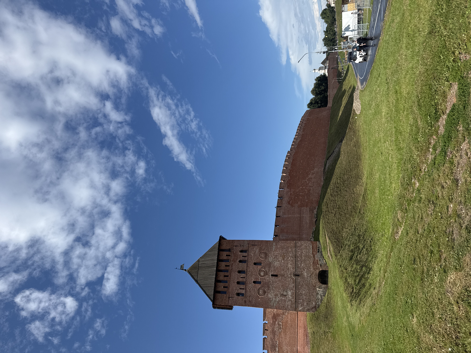 This is a daytime, outdoor, low-angle shot of the Veliky Novgorod Kremlin, also known as the Novgorod Detinets. The main subject is a tall, red brick tower, situated on a grassy embankment. The tower has a dark, pyramidal roof and multiple rows of small, arched windows. The wall of the Kremlin, also made of red brick, extends from the tower to the right, curving around the landscape. The sky is a vibrant blue with scattered white clouds. In the background, a paved road with two people walking and a white car is visible, along with trees and distant buildings, including a white church with a dome. The overall scene depicts a historic fortress on a sunny day.
