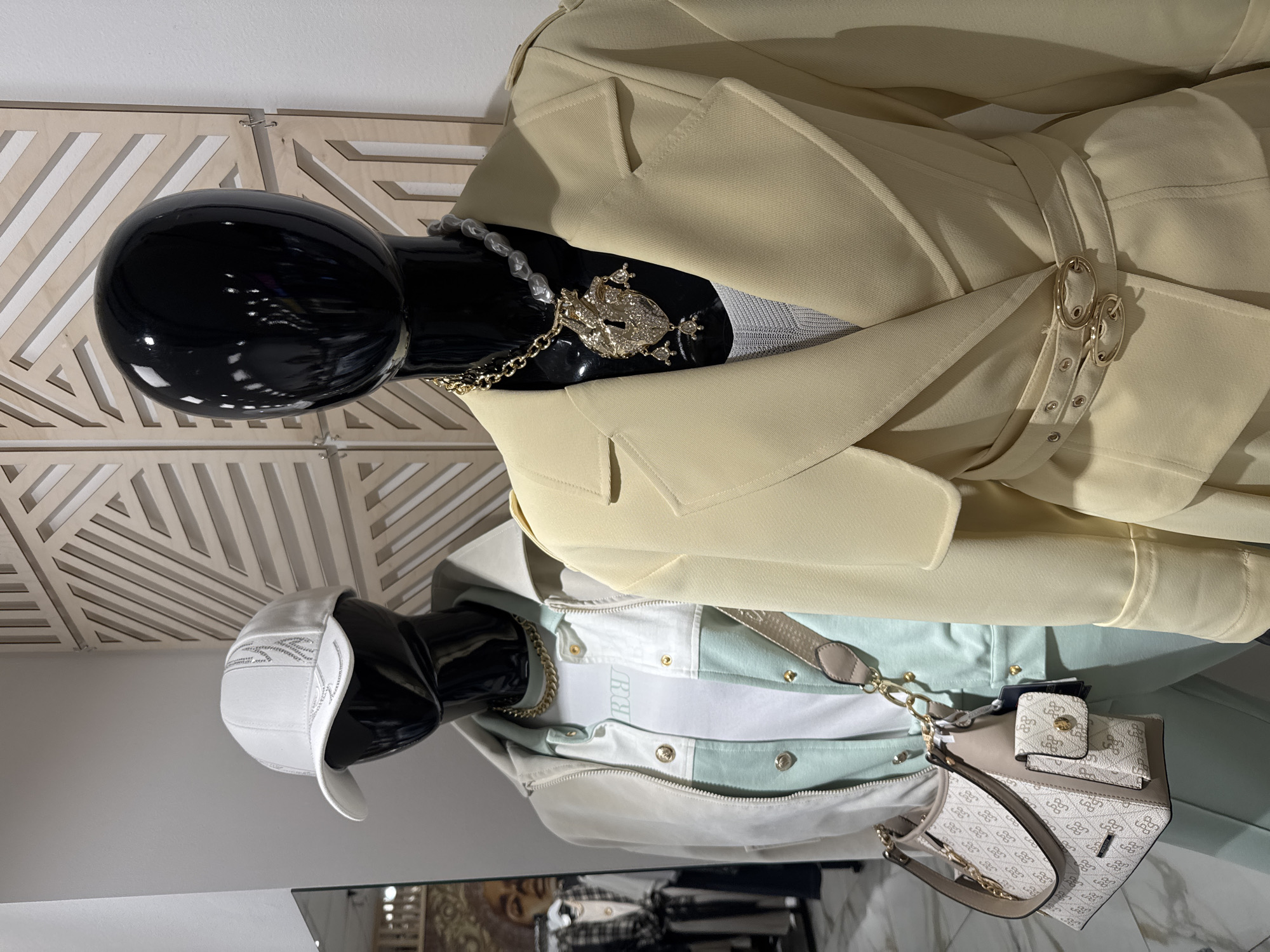 The image shows two mannequins in a retail setting. The mannequins are black and feature clothing ensembles. The top mannequin wears a light tan trench coat and a large gold frog pendant necklace with pearl accents. The bottom mannequin wears a white and light green jacket with a white baseball cap and a small handbag with a chain strap. The background includes a light-colored wooden wall decoration with geometric cutouts. The scene likely takes place in a retail store in Saint Petersburg, Russia.