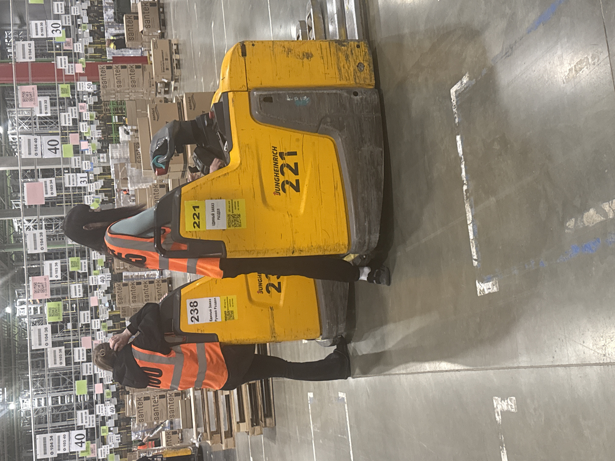 The image depicts a warehouse scene, likely in an industrial area, possibly in Lupanovo, Russia. Two people wearing orange safety vests and dark clothing are standing in front of two yellow Jungheinrich electric pallet jacks, labeled "238" and "221." The jacks appear to be in a warehouse aisle filled with stacked goods, many with "Santek" branding. The aisle is structured with numbered shelves and numerous QR codes. The floor is a grey industrial surface with white and blue line markings. The overall impression is one of a busy distribution center.