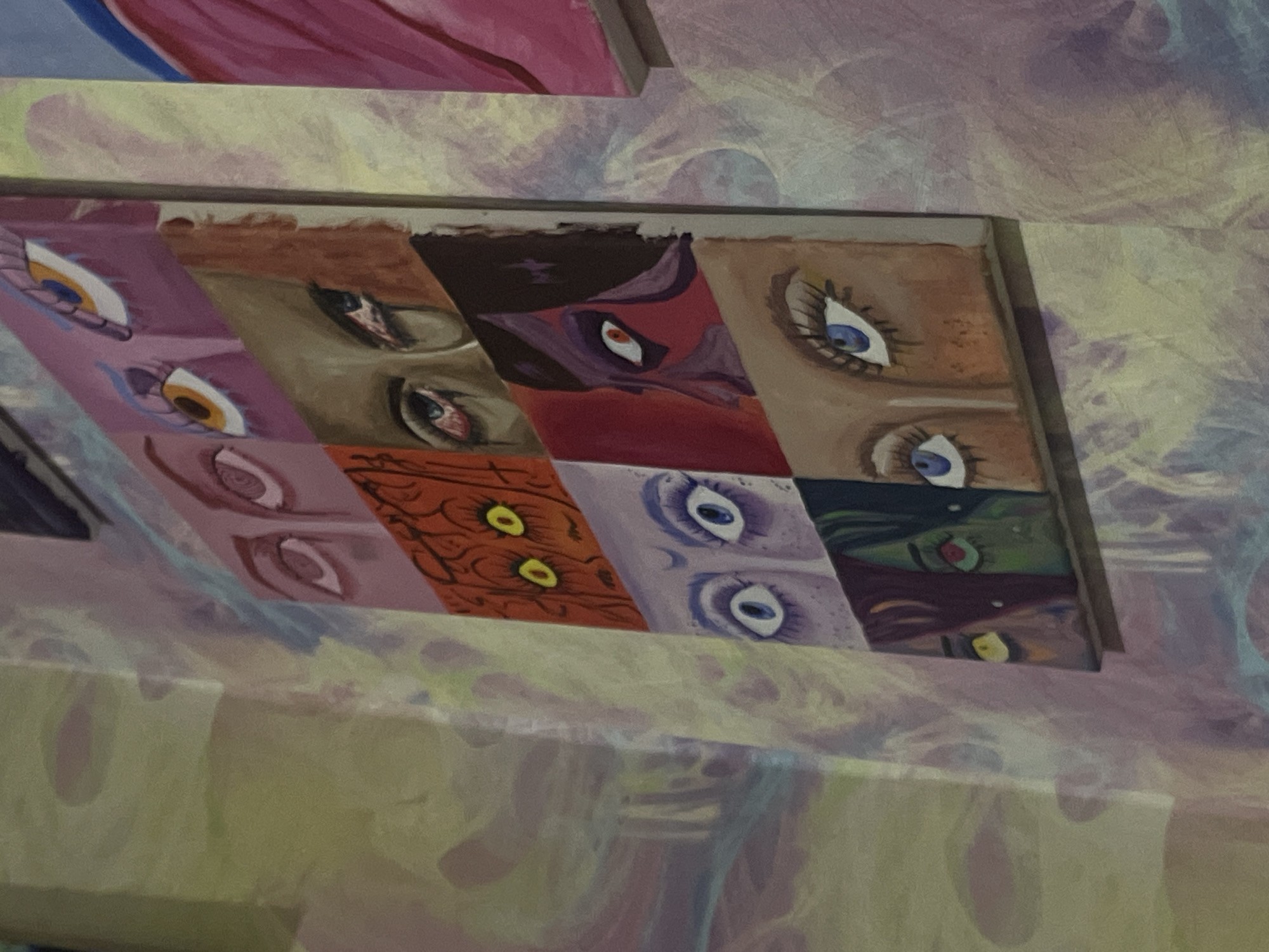 The image shows a close-up of a vertical, multi-panel painting of eyes, affixed to a wall with a patterned wallpaper. The painting is divided into nine rectangular sections, each featuring a different pair of eyes or a single eye. The styles and colors of the eyes vary widely, suggesting a diverse range of subjects and artistic interpretations. The wallpaper behind the painting features a swirling, abstract pattern in pastel shades of yellow, pink, and purple. To the right of the main painting, another canvas with pink and blue abstract elements is partially visible. The lighting in the image appears to be ambient, creating soft shadows and highlighting the textured surface of the wallpaper and the painting. The overall impression is of an art installation or display in an indoor setting. The location provided, "ЖК «Борисоглебское», Russia," suggests this artwork is situated within a residential complex in Borissoglebsk, Russia.