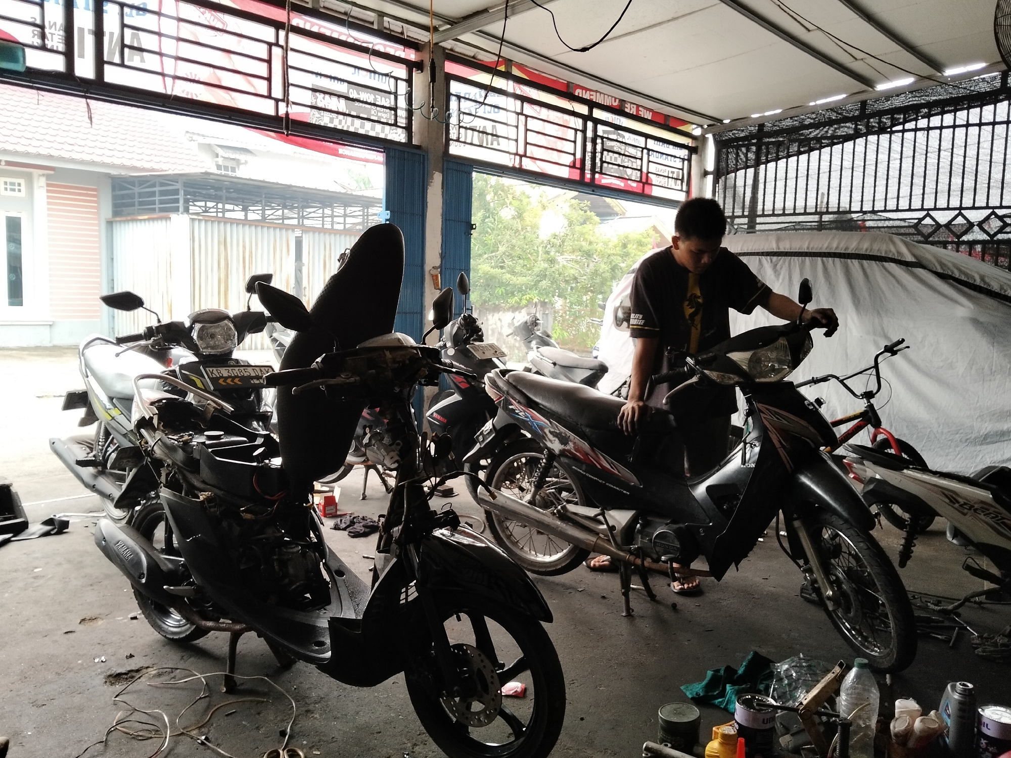 The image depicts an outdoor workshop or repair area, likely located in Arang Limbung, Indonesia, as suggested by the context. The scene is filled with motorcycles, some of which appear to be in various states of repair. In the foreground, a dark-colored scooter is prominently featured, its engine compartment exposed, revealing internal mechanics and wiring. To the right, another scooter is partially visible, with a person in a black t-shirt standing next to it, appearing to be working on it. The background shows more motorcycles, a white tarp covering a larger object, and the open doors of a building leading to an outdoor area with trees. Various tools and spare parts are scattered on the ground, indicating an active repair environment. The overall impression is one of a functional, albeit somewhat cluttered, motorcycle repair shop.