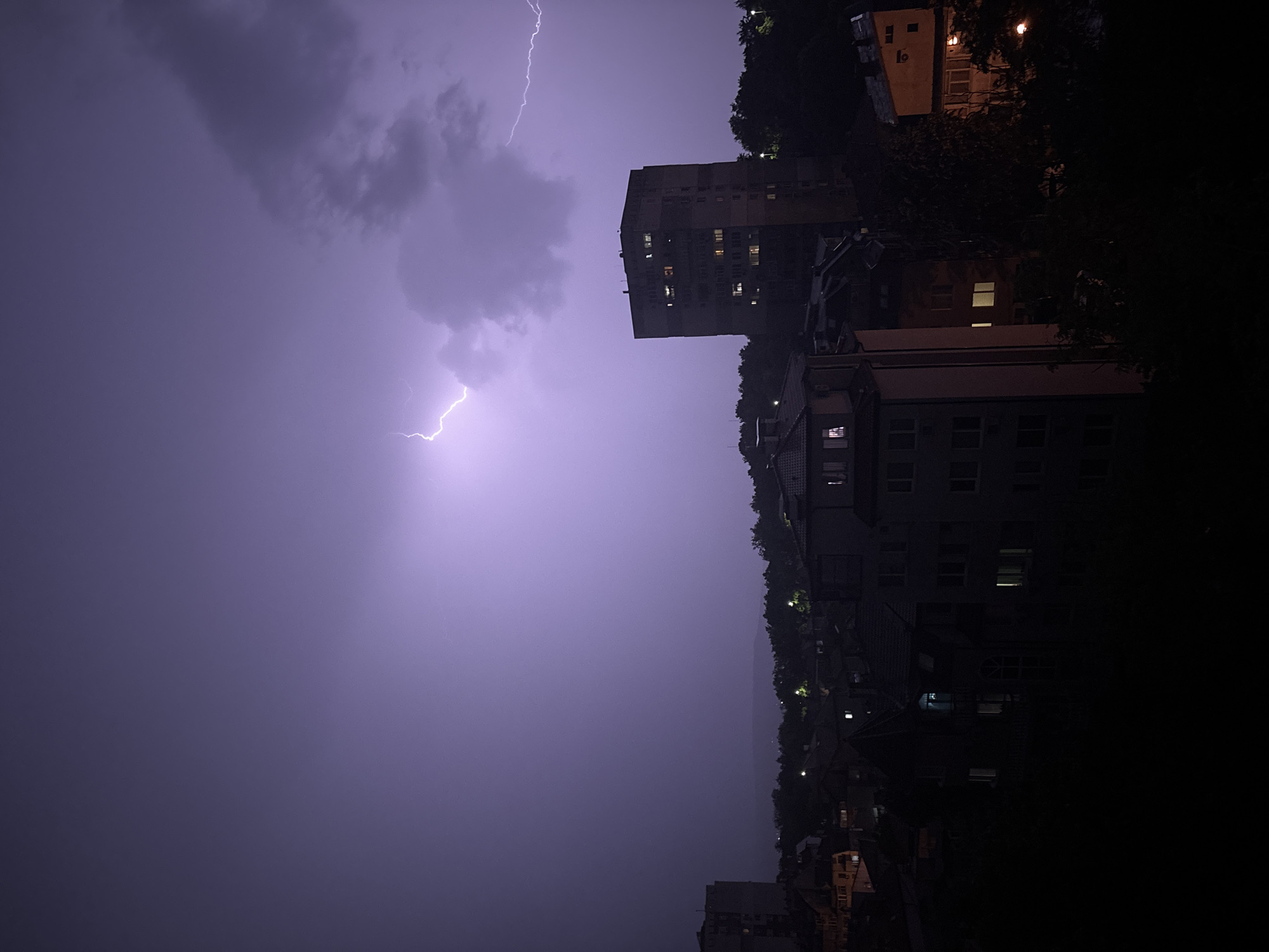 The image depicts an urban landscape at night during a thunderstorm, illuminated by lightning, in Varna, Bulgaria. The sky is largely covered by dark clouds, intensely illuminated by a prominent, branching lightning strike in the upper left-center, and another diffuse, multi-pronged lightning bolt extending from the upper right. The lightning casts a vivid purple and lavender hue across the sky. In the midground, a tall, multi-story concrete apartment building is centrally positioned on the right side of the frame, its facade revealing numerous windows, several of which are internally illuminated. To the left and below this structure, and extending across the midground, are various other residential buildings, mostly dark, with occasional illuminated windows. Dense, dark tree canopy partially obscures the lower portions of buildings and fills the spaces between them. No individuals are visible, but the illuminated windows imply human presence within the structures. The immediate foreground is dark and undefined.