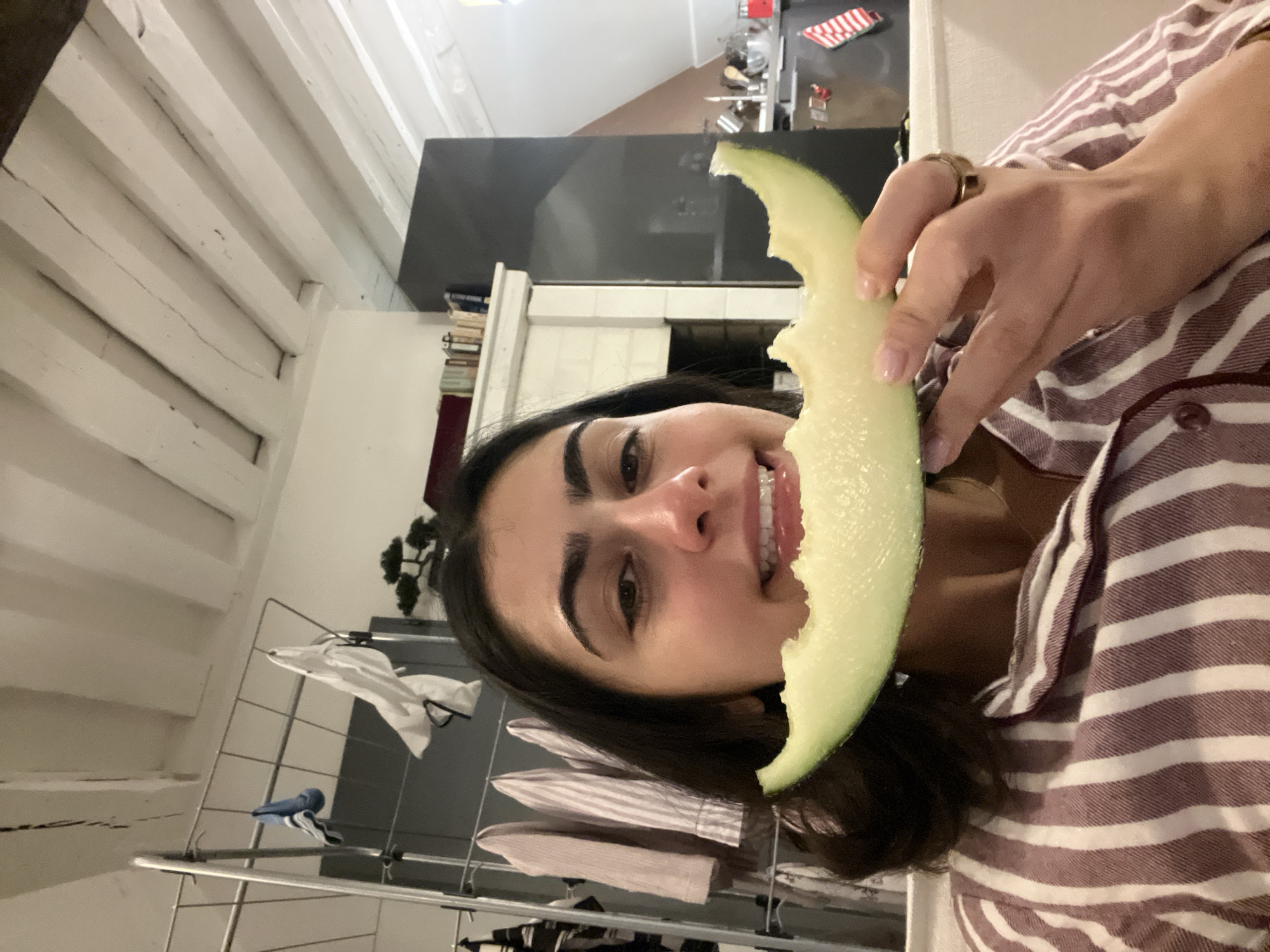 A woman is holding a slice of honeydew melon up to her face, seemingly about to take a bite. She is wearing a striped shirt. Behind her is a drying rack with clothes on it. The background suggests the interior of an apartment, possibly in Paris, with exposed beams in the ceiling and a modern kitchen visible in the distance.