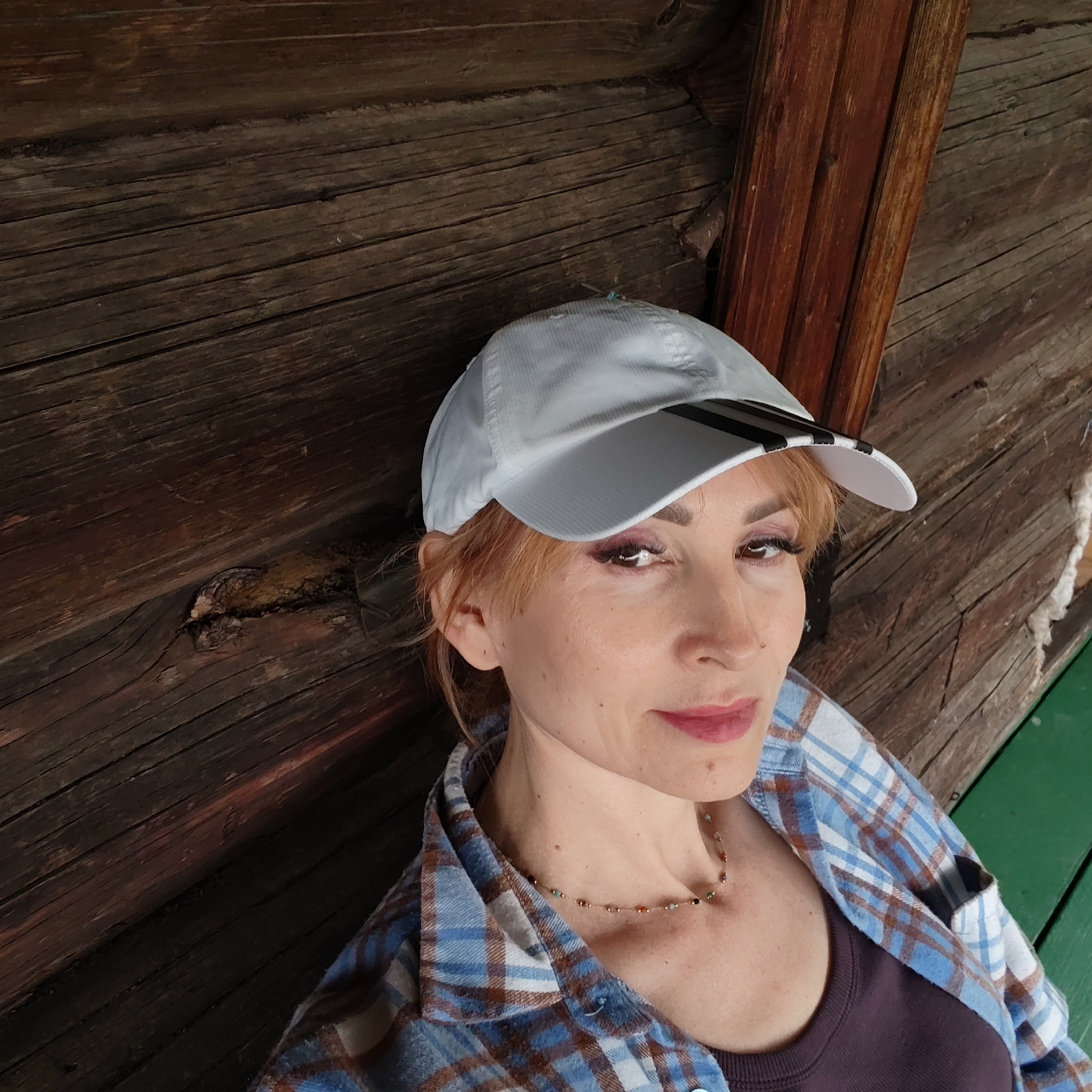 The image depicts a woman wearing a white baseball cap with black stripes on the bill, a blue and brown plaid flannel shirt over a dark purple top, and a delicate beaded necklace. She has red hair and is wearing makeup. The background consists of dark brown wooden planks, possibly part of a rustic building or fence. A dark red wooden beam is also visible. The woman is leaning against the wooden surface, suggesting a relaxed or casual pose. The location in the city of Chaikovsky, Russia, is not discernible from the visual information in the image.