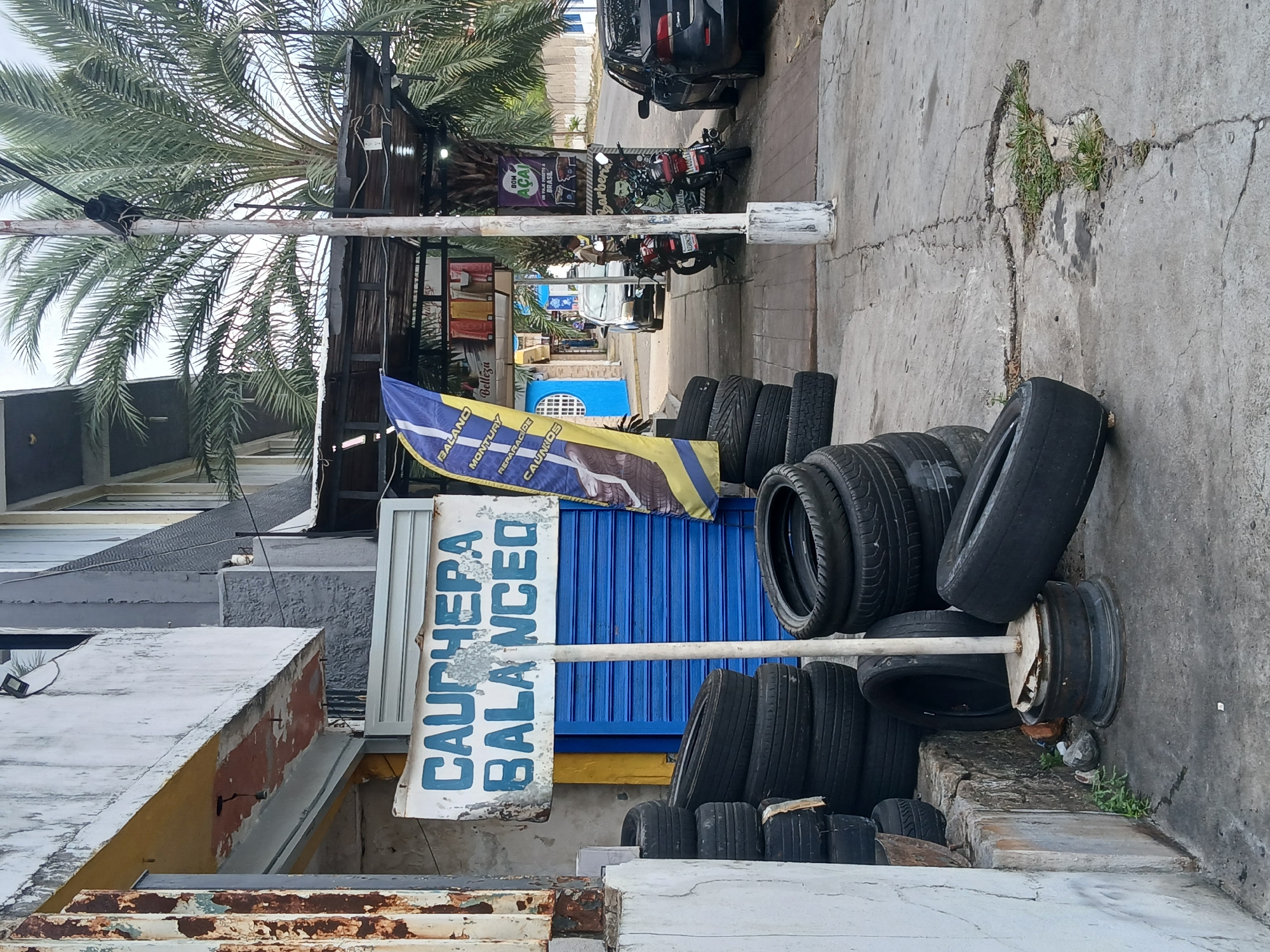 The image depicts a street scene in Guayana City, Venezuela, showing a tire shop. Several stacks of used tires are stacked on the sidewalk next to a sign that reads "CAUCHERA BALANCED." The tires are of various sizes and conditions. A flag is also visible, partly obscuring the shopfront. In the background, there are other businesses, parked motorcycles, and cars along the street. The street is paved with concrete and has some cracks and weeds growing. A palm tree is visible.