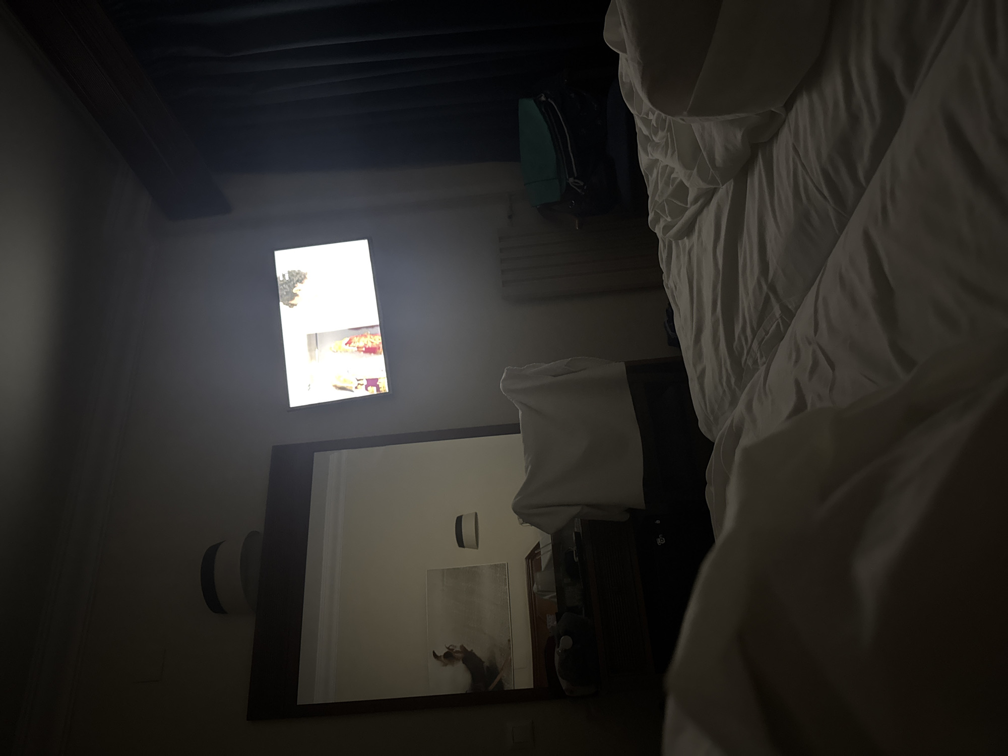 The image depicts a dimly lit hotel room with a large unmade bed in the foreground. To the left, a large mirror reflects part of the room, showing a desk with items on it and a piece of artwork on the wall. A white chair draped with fabric is visible in the middle ground, positioned near the mirror. A television screen is on the wall to the right, displaying a bright image. A dark blue curtain is drawn on the far right side of the room, and a white radiator or heating unit is situated below the television. A dark-colored suitcase and a teal bag are placed on a chair to the right of the radiator. The overall impression is of a lived-in space, likely a hotel room in Fuengirola, Spain, given the context provided.