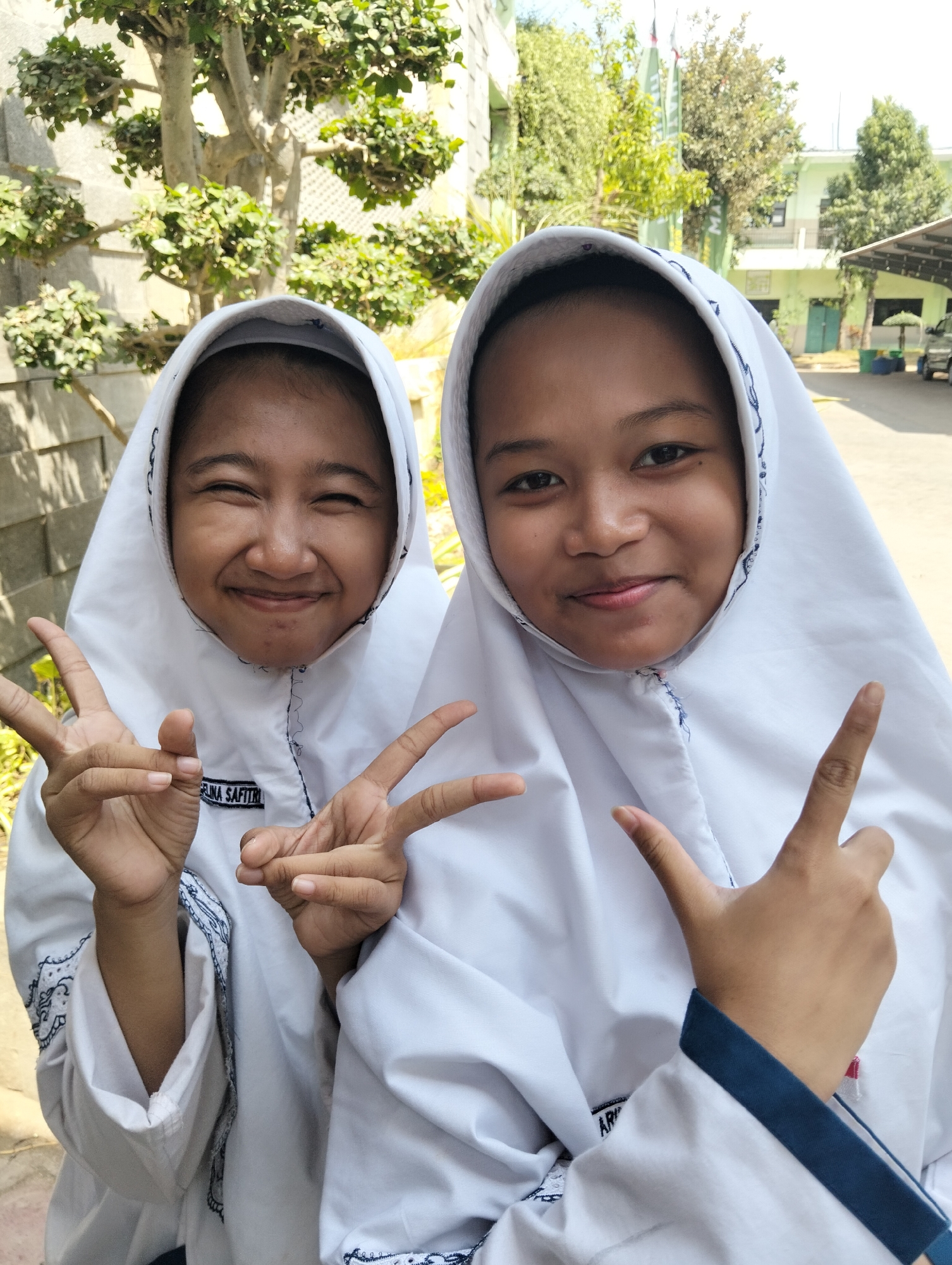 Two young Indonesian girls, appearing to be school-aged and wearing white hijabs and uniforms, pose for a photograph outdoors. The girl on the left is smiling broadly with her eyes closed and is making a peace sign with her left hand, while her right hand is also raised in a peace sign gesture. The girl on the right is smiling gently and making a peace sign with her right hand, her left hand is also raised. Both girls have name tags on their uniforms; the one visible on the left girl's uniform reads "SELINA SAFITRI". They are standing in what appears to be a schoolyard or outdoor area with trees and some buildings in the background. The environment suggests a warm, sunny day. The location is the yard of a school in Paciran, Indonesia, as indicated by the presence of a building with a green roof and a white wall, characteristic of many educational institutions in the region.