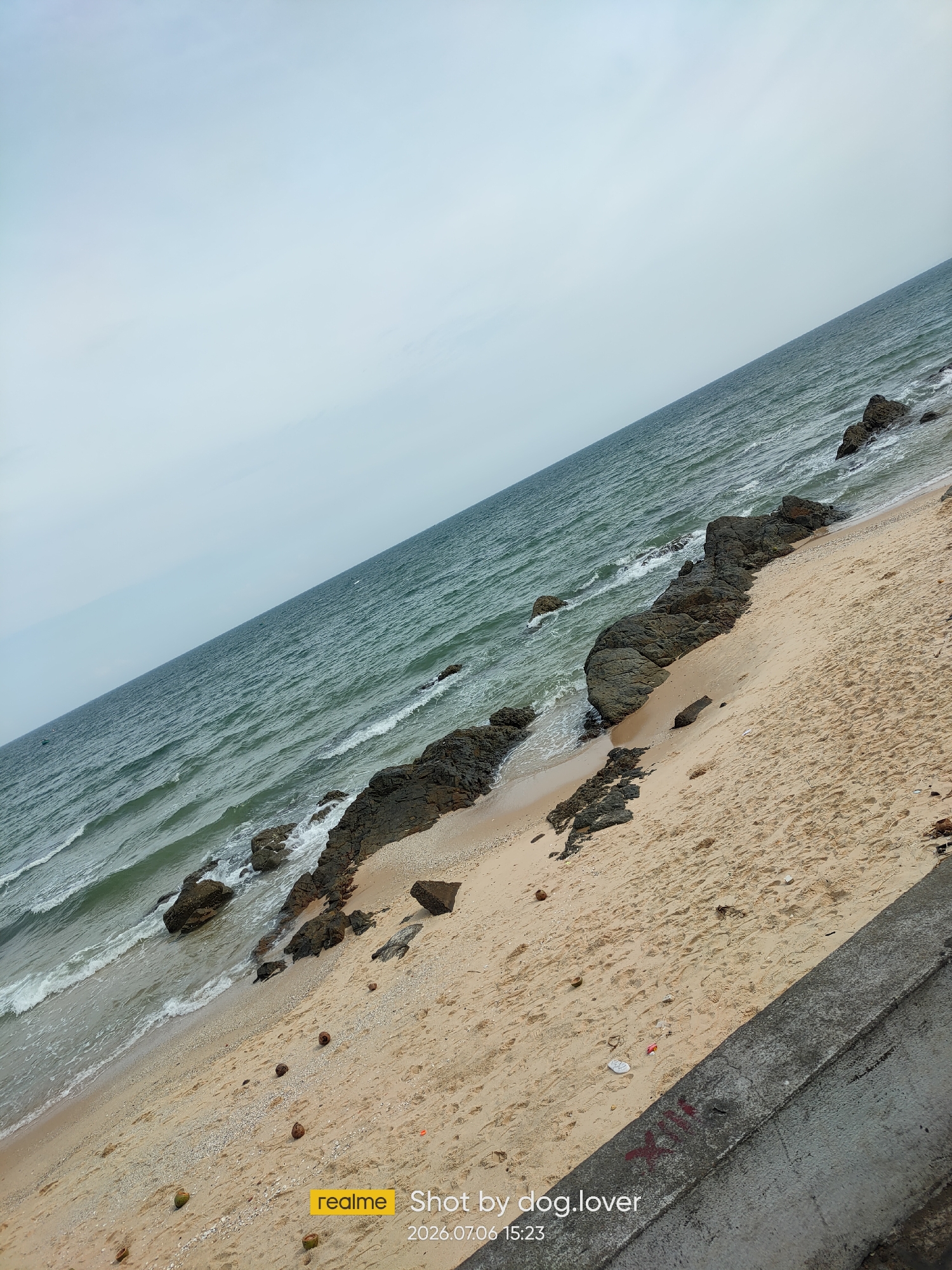 The image displays a diagonal view of a sandy beach with scattered rocks and the ocean. The sky is overcast with a pale blue hue. Waves gently lap against the shore and the rocky formations. The sand is light brown and appears to have footprints and a few scattered coconut shells. In the foreground, a concrete barrier runs along the edge of the beach. A watermark in the bottom right corner indicates "realme Shot by dog.lover 2026.07.06 15:23."