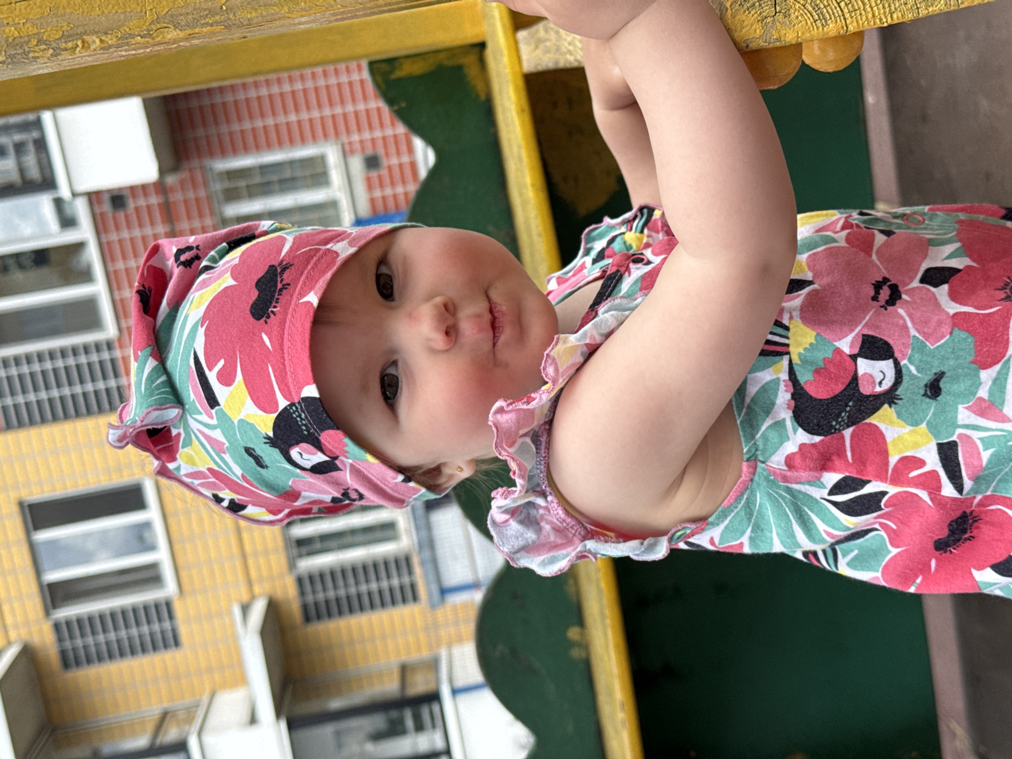 A close-up, eye-level outdoor medium shot captures a toddler girl wearing a colorful floral print outfit and matching headscarf. She is positioned on a playground structure, with a yellow support post partially obscuring her right side. The girl faces the camera, her gaze direct, with a slight pucker of her lips. Her right arm is visible, resting on the post. In the background, out-of-focus, is a building with yellow and brick-red facades, featuring multiple windows and balconies. The playground equipment appears to be painted green and yellow. The scene is situated in an urban environment.