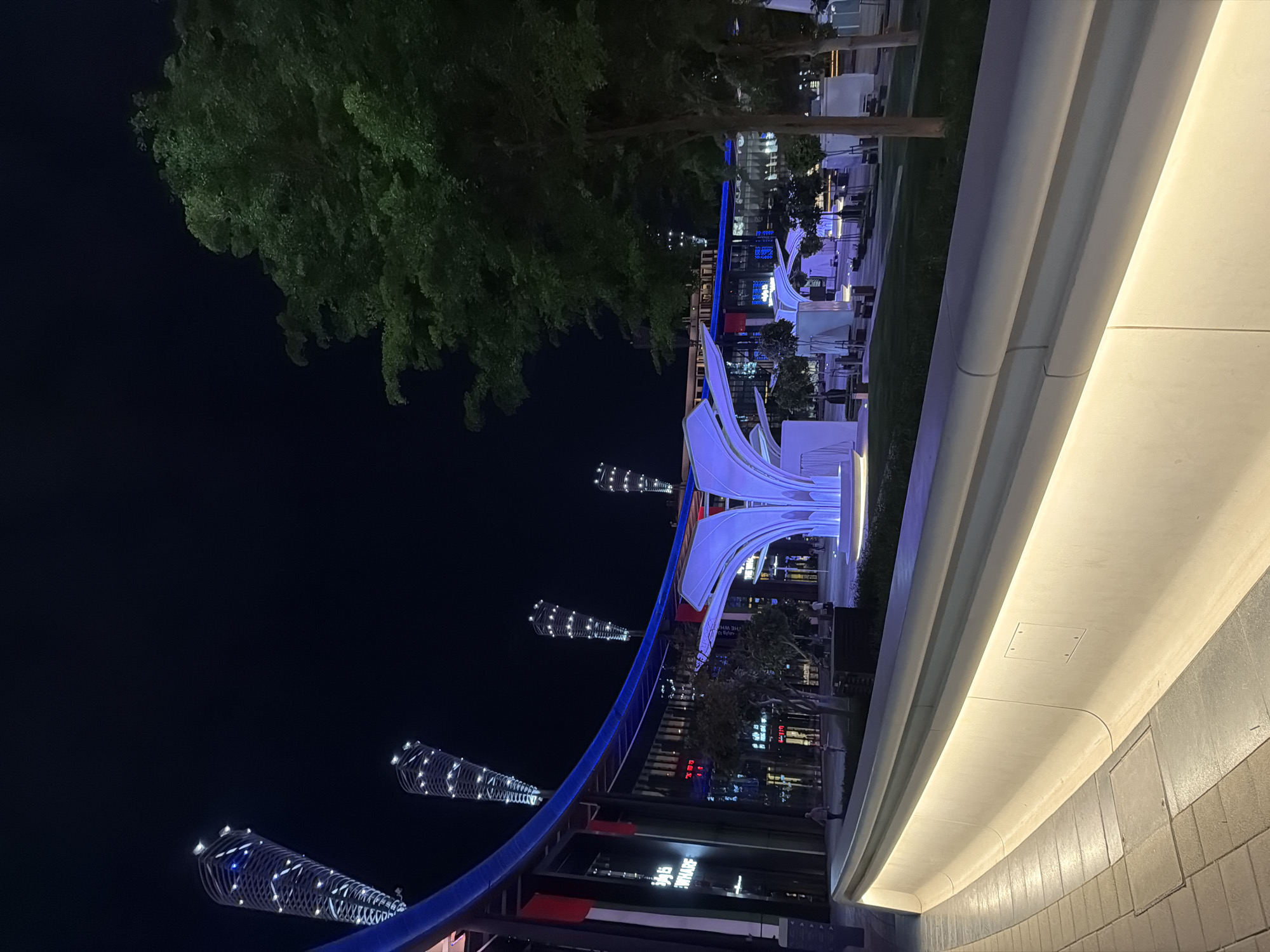 The image displays a modern outdoor public space at night, likely a promenade or entertainment area in a city like Dubai, UAE. The scene is dominated by artificial lighting, creating a vibrant and contemporary atmosphere.
In the midground, a prominent, white, abstract sculpture with flowing, curved forms is bathed in cool blue and purple light, serving as a central illuminated feature. A long, dark blue, curved structural element acts as a visual guide, running through the mid-left portion of the frame. To the left, several tall, decorative conical or spiral-shaped structures, intricately illuminated with white lights, rise against the dark night sky.
The right side of the foreground is occupied by a substantial, light-colored, curved wall or barrier, which is internally lit, casting a warm, soft glow downwards. Beyond this, and in the background, are outdoor seating areas with tables and chairs, suggesting cafes or restaurants. Green trees with visible foliage are scattered throughout the landscape, some partially illuminated. Modern buildings with illuminated windows and facades are visible in the further background. On one building, a sign displaying Arabic script followed by the English word "SQUARE" can be seen.
The time of day is clearly night, with a dark sky and the entire area illuminated by various light sources. There are a few indistinct human figures visible in the distance, suggesting people are present, likely enjoying the evening ambiance. The overall impression is one of sophisticated urban design and a lively nighttime destination.
