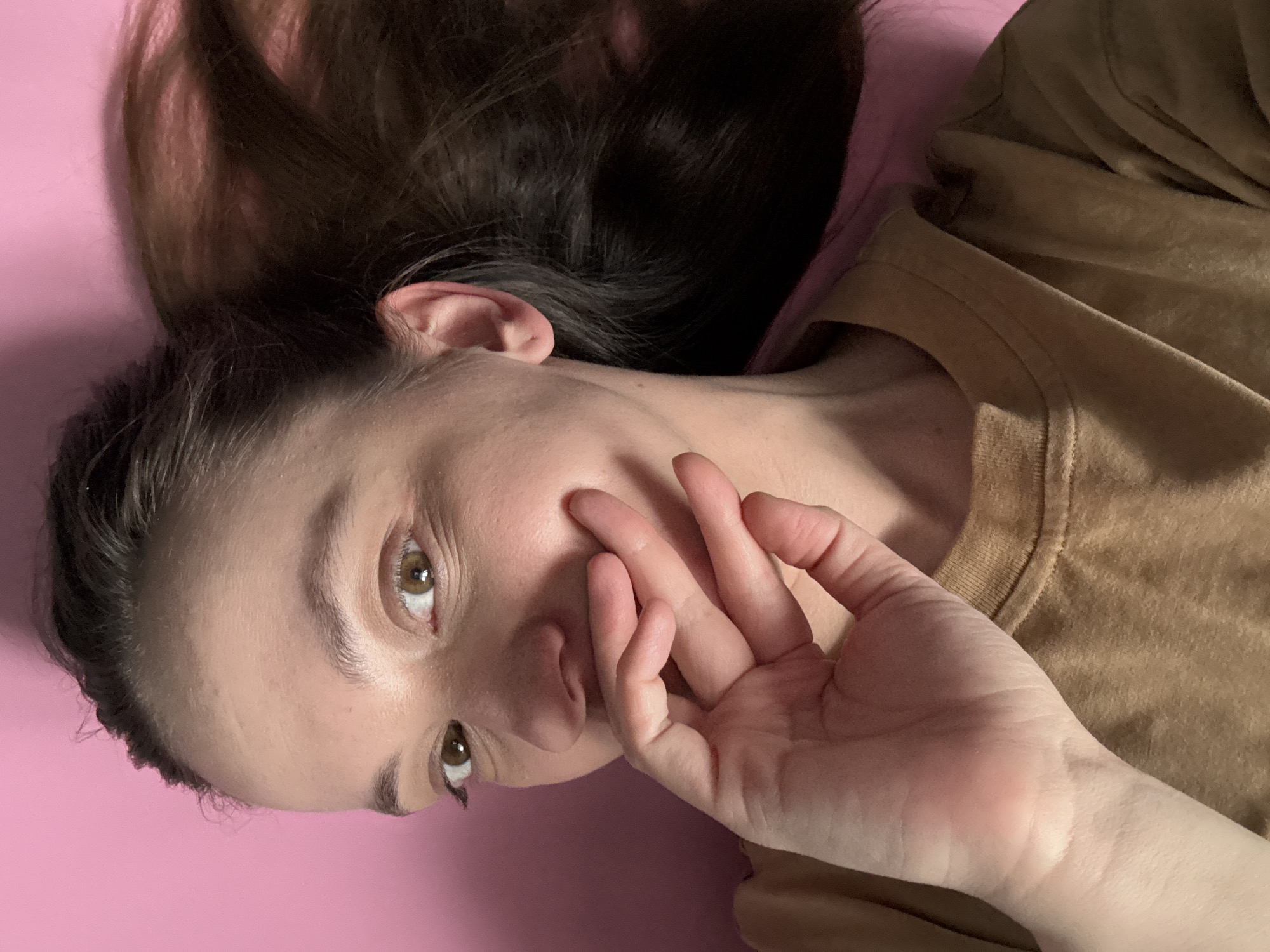 A young woman with long, dark brown hair is lying on a pink surface. She is wearing a tan or light brown shirt. Her head is tilted to her left, and her right hand is raised near her face, fingers slightly curled. Her eyes are open and appear to be brown. The background is plain and pink. There is no evidence to suggest the location of the image is in Chaikovsky, Russia.
