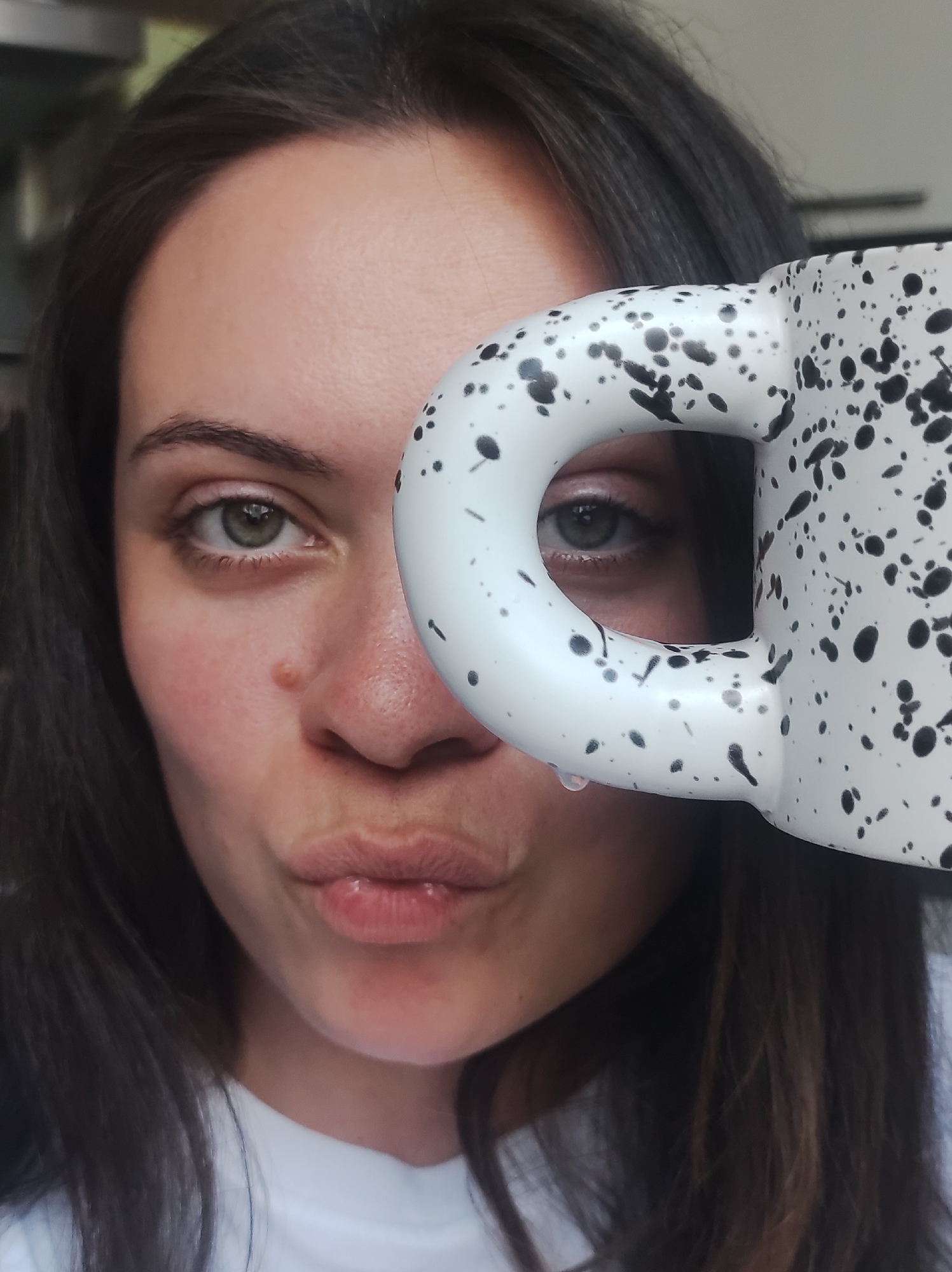 This is a close-up, medium shot of a woman's face, partially obscured by a white mug with black speckles. The woman has dark hair and green eyes, and she is making a puckered-lip expression. A single drop of liquid is visible on the rim of the mug, near her mouth. The background is blurred and out of focus, but appears to be an indoor setting. The specific location in Chaikovsky, Russia is not discernible from the image.