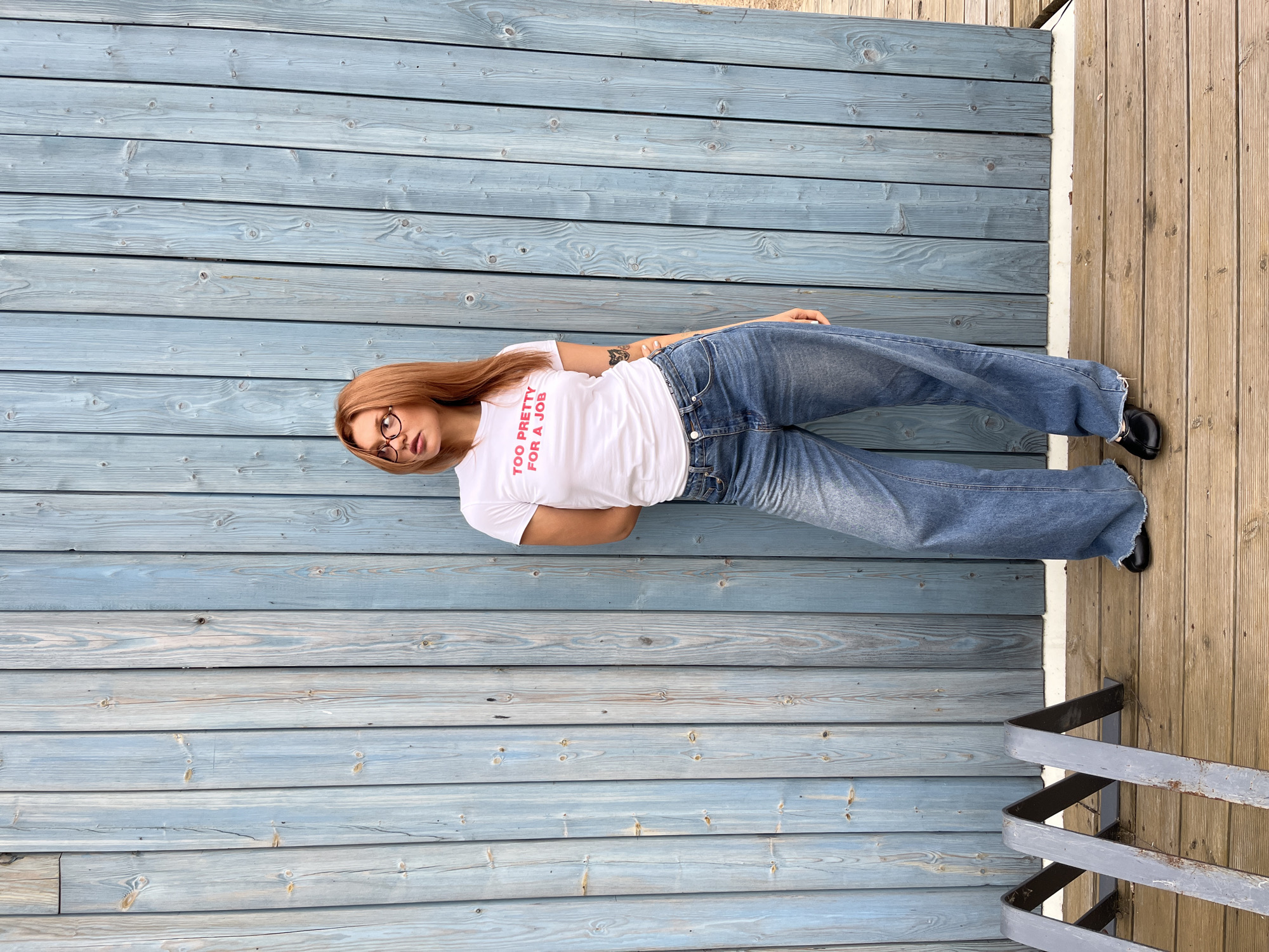 A full-length outdoor portrait captures a woman standing against a vertical wooden plank wall painted in a desaturated blue-gray hue, located in Chaikovsky, Russia. The woman possesses long, reddish-blonde hair and wears reddish-brown framed eyeglasses. She is dressed in a white short-sleeved t-shirt displaying the text "TOO PRETTY FOR A JOB" in pink on the chest. She wears wide-leg, light blue denim jeans with frayed hems, beneath which a white waistband with faint "Calvin Klein" text is partially visible. Her footwear consists of black flat shoes with a metallic embellishment on the vamp. A small tattoo, consistent with a butterfly design, is present on her upper left arm. The woman stands with her head turned to her right, looking upwards and to her left. The ground consists of light brown wooden decking. To the left foreground, a multi-sectioned metal bike rack, dark gray and showing signs of rust, is visible.
