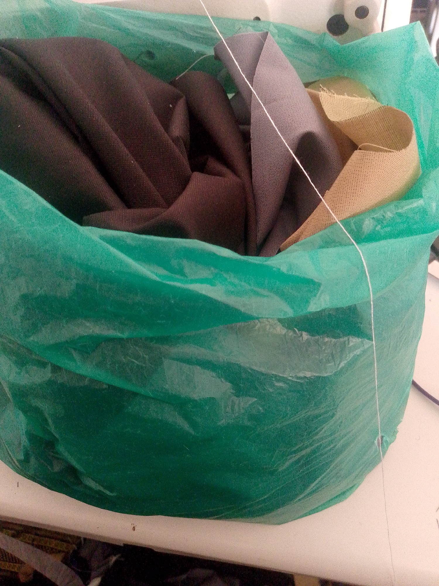 A medium shot captures a large, crinkled green plastic bag filled with assorted fabric scraps. The bag is open at the top, revealing folded pieces of dark brown, grey, and tan fabric. A thin white thread or string runs across the fabric and out of the bag, suggesting it might be part of a sewing project. The bag rests on a light-colored, possibly white, surface. In the background, a blurred white object with black circles is visible, resembling a sewing machine head. The overall scene is domestic or workshop-related, with no discernible connection to Jalingo, Nigeria.