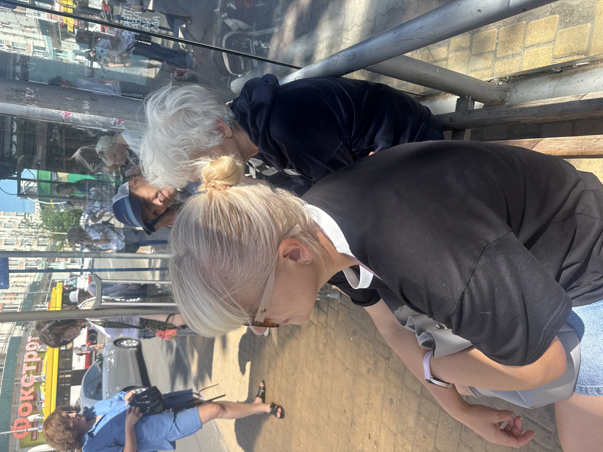 In Odesa, Ukraine, at a bus stop, several individuals are captured in a candid moment. The foreground is dominated by a woman with blonde hair pulled back in a bun, wearing sunglasses and a black t-shirt with white trim. To her right, another person with white hair is seated, wearing a dark blue hooded sweatshirt. Behind them, a man wearing a blue baseball cap and a dark t-shirt is visible. Further in the background, a woman in a blue shirt and shorts stands, holding a black bag. A yellow bus with Russian text is seen on the street, suggesting a busy urban environment. The scene is illuminated by bright sunlight, casting shadows on the brick sidewalk and the glass panels of the bus stop.