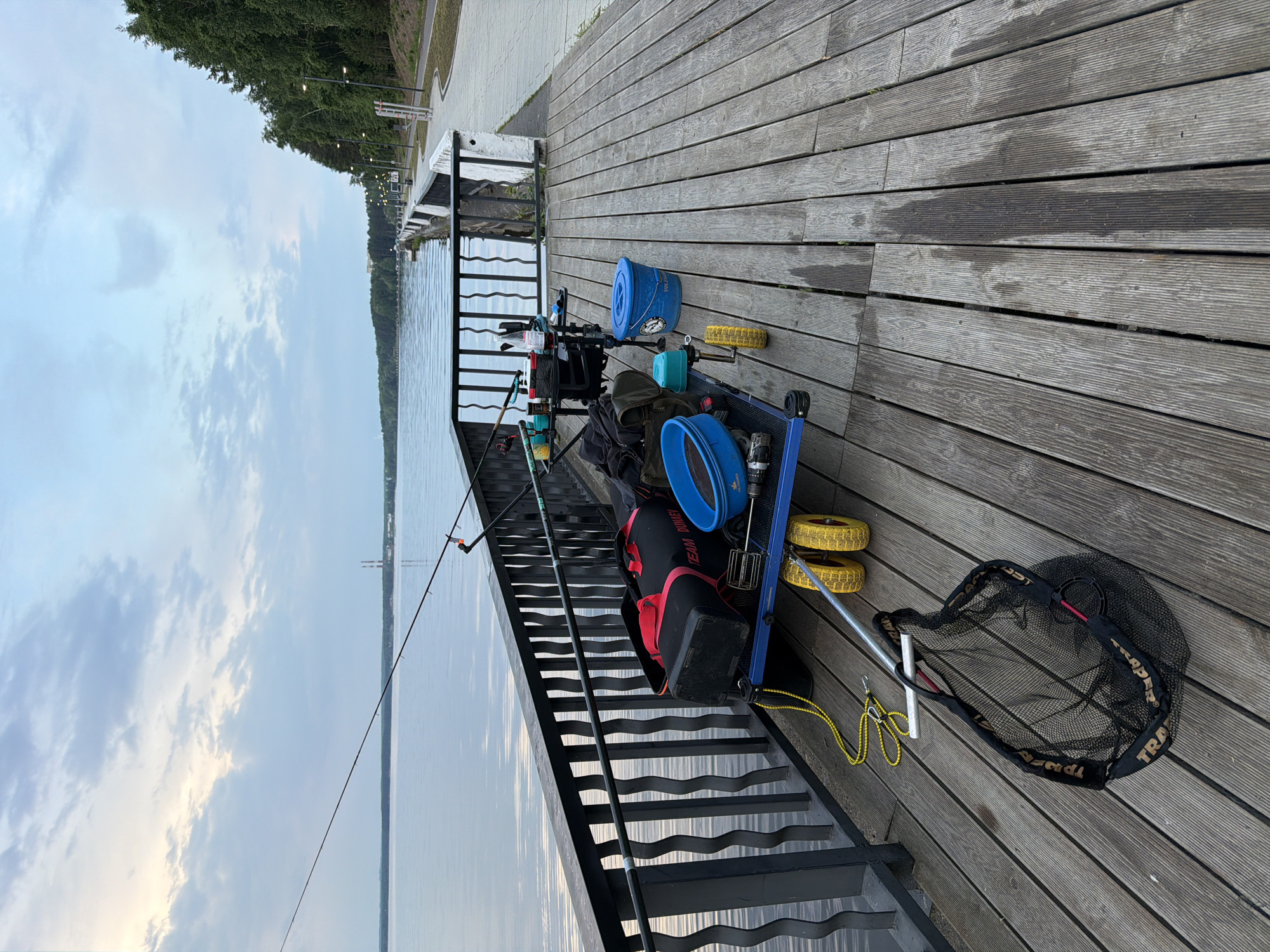 The image captures a detailed setup for fishing on a wooden pier next to a calm body of water, likely a river or lake, in Chaikovsky, Russia. The scene is presented from a slightly elevated perspective, looking down towards the water.
In the foreground and midground, a blue fishing trolley is heavily loaded with various fishing equipment. A prominent black and red bag, bearing the text "TEAM DUNAFF," sits on the trolley alongside multiple blue buckets or bowls, a turquoise container, and other smaller bags and gear. The trolley itself has distinct yellow, possibly foam, wheels. Resting beside the trolley is a large black landing net, clearly displaying the brand "TRAPER" on its rim. Two fishing rods are visible; one extends over the water from a rod rest, while another rests against the trolley and pier railings. A coiled yellow rope with a carabiner is also visible near the landing net.
The pier itself is made of weathered grey wooden planks, some of which appear wet or stained, suggesting recent activity or weather. To the left, metal stairs with an open risers lead down from the pier to the water's edge.
The background features a vast expanse of calm, grey-blue water, reflecting the sky. On the opposite bank and further along the pier, a dense line of green trees is visible, with a paved pathway and streetlights running parallel to the water's edge. The sky above is cloudy, with soft, diffused light, suggesting either early morning or late afternoon. No individuals are present, but the arrangement of the specialized equipment strongly indicates preparation for or engagement in coarse or feeder fishing. The overall atmosphere is tranquil and focused on the angling activity.