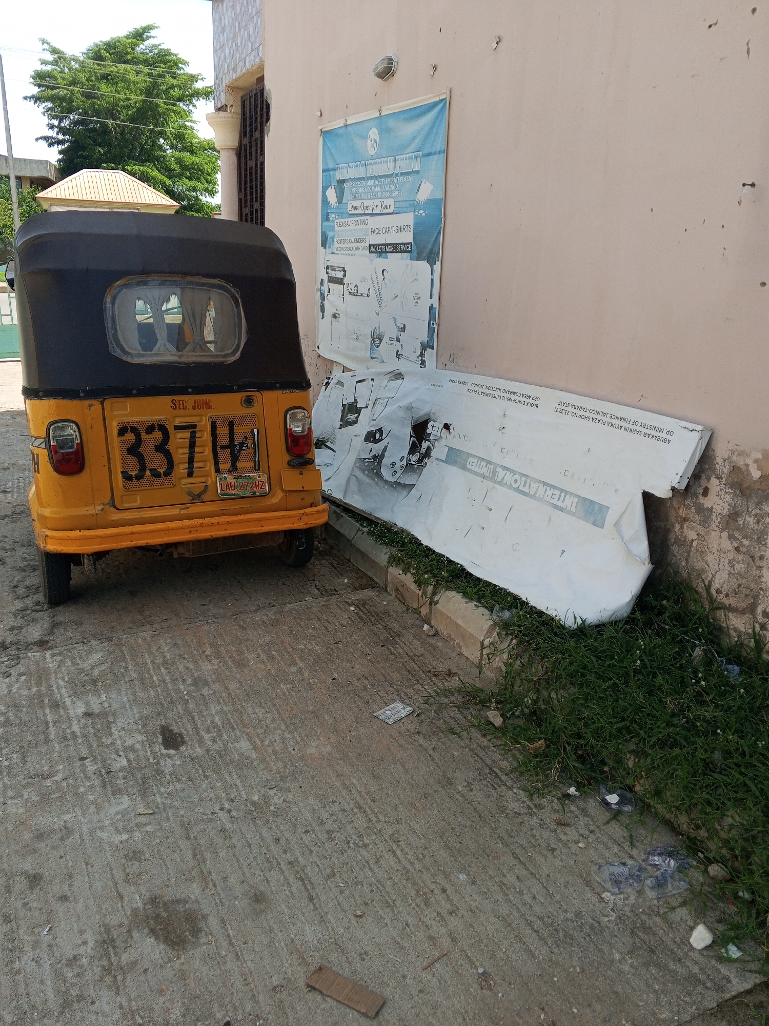 An outdoor scene depicts a yellow and black auto-rickshaw parked on a paved surface, likely a street or alley in Jalingo, Nigeria. The auto-rickshaw, which appears to be a Keke Napep, is facing to the left and its rear license plate, displaying "LAU 272 MZ", is clearly visible. Above the license plate, a larger number "337H" is stenciled onto the vehicle. To the right of the auto-rickshaw, a large, white, tattered banner lies on the ground, partially covered by grass and debris. The banner has text printed on it, including "INTERNATIONAL LIMITED" and what appears to be a list of addresses and contact information. The wall behind the banner is light-colored and shows signs of age and wear. In the background, lush green trees are visible, suggesting a tropical or sub-tropical environment. The overall impression is one of a somewhat neglected urban setting.