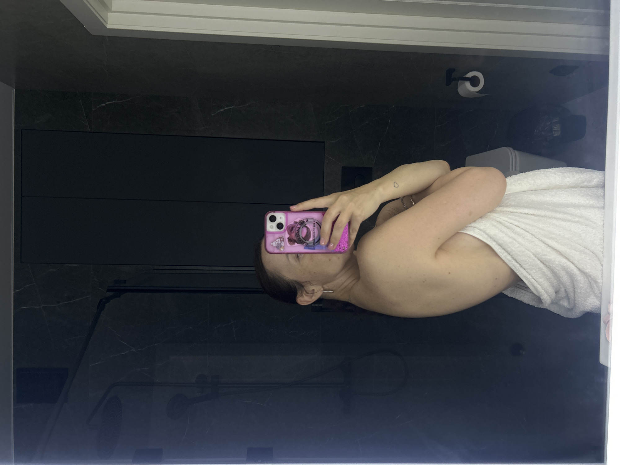 A woman is depicted taking a mirror selfie in a modern bathroom. She is wrapped in a white towel, covering her lower body while her back and right shoulder are exposed. Her face is largely obscured by a pink smartphone held in her right hand, which features a glittery pink case adorned with a bow and reversed text that includes "ORIGINAL." A delicate, elongated earring dangles from her left ear, and a small heart-shaped tattoo is visible on her left bicep.
The bathroom interior, visible in the mirror's reflection, features dark grey or black marble-patterned wall tiles. To the left, a shower enclosure is equipped with a dark-finished rain showerhead and a handheld shower fixture. Above a white toilet, positioned to the right of the woman's reflection, are dark grey, possibly black, wall-mounted cabinets. A black toilet paper holder with a white roll is mounted on the wall to the right of the toilet. A white door frame is partially visible on the far right edge of the frame.
Based on the visual information within the image, there are no identifiable architectural features, signage, or specific environmental cues that indicate the location is Yekaterinburg, Russia.