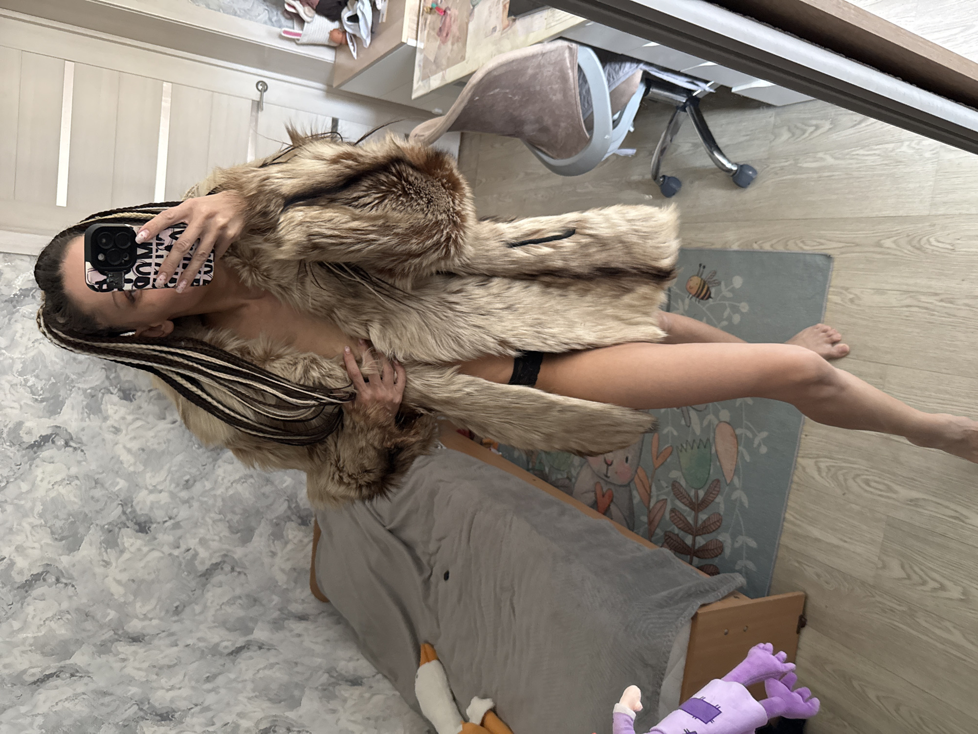 A woman with dark hair and blonde streaks, wearing a large fur coat and black underwear, stands in front of a mirror in an indoor environment. She is holding a smartphone with a patterned case, seemingly taking a selfie. The floor is light-colored wood, partly covered by a blue rug with illustrations. A bed with a grey blanket and various plush toys is visible. The setting appears to be a bedroom or living space within a domestic residence. The location is not discernible as being in Bor, Russia based on visual details.