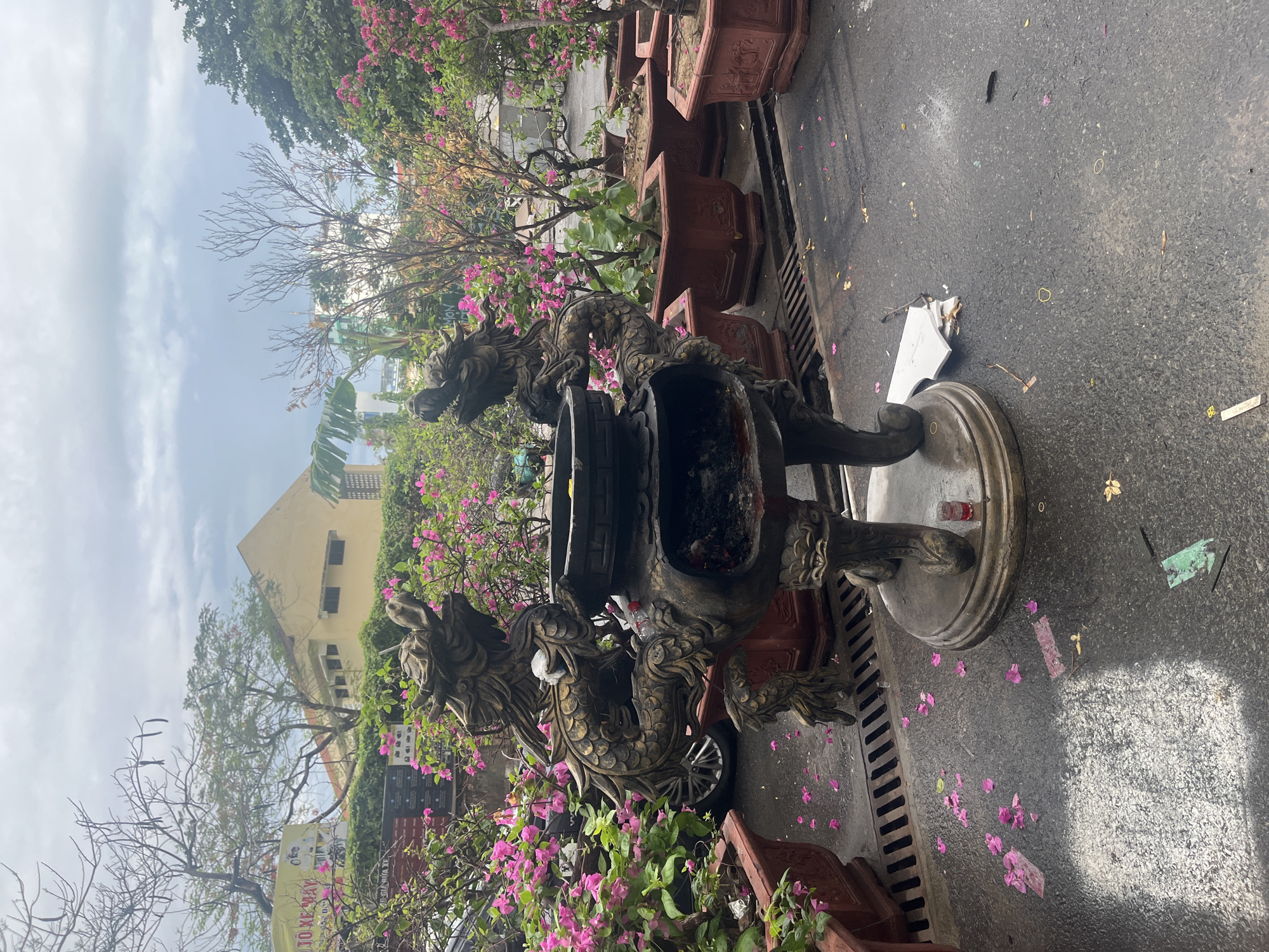 This outdoor scene shows an ornate incense burner with dragon sculptures on a paved surface in Bac Nha Trang Ward, Vietnam. The burner is surrounded by potted plants with pink flowers. The background includes buildings, trees, and an overcast sky. Litter and flower petals are scattered on the ground.