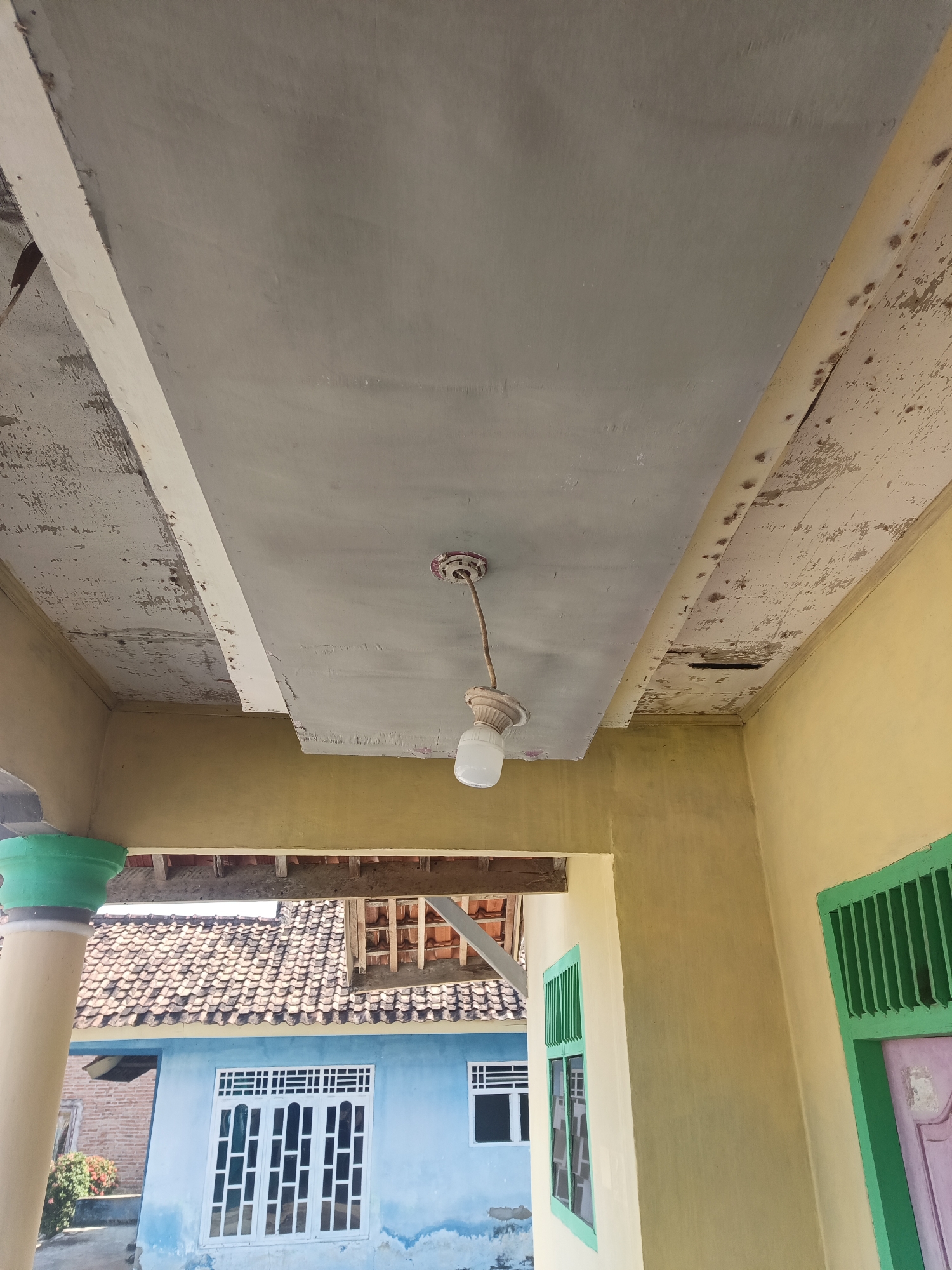 The image captures an upward view of an exterior ceiling structure and surrounding buildings. The ceiling itself appears to be constructed of a grey panel with a lighter colored trim, showing signs of wear and peeling paint, particularly on the left side. A light fixture with a white bulb hangs from the center of the ceiling panel. To the right of the ceiling, a yellow wall is visible, also exhibiting some discoloration and peeling paint. Below the ceiling, a section of a blue house with white-framed windows is in view, with a terracotta tiled roof above it. Further to the right, another section of the yellow building is present, with a green-framed window and a pink door partially visible. The image is taken from a low angle, looking up towards the ceiling and the buildings in the background. The overall impression is of an outdoor area, possibly a porch or walkway, attached to a residential building in what appears to be a rural or suburban setting in Terbanggi Marga, Indonesia. There are no people visible in the image.