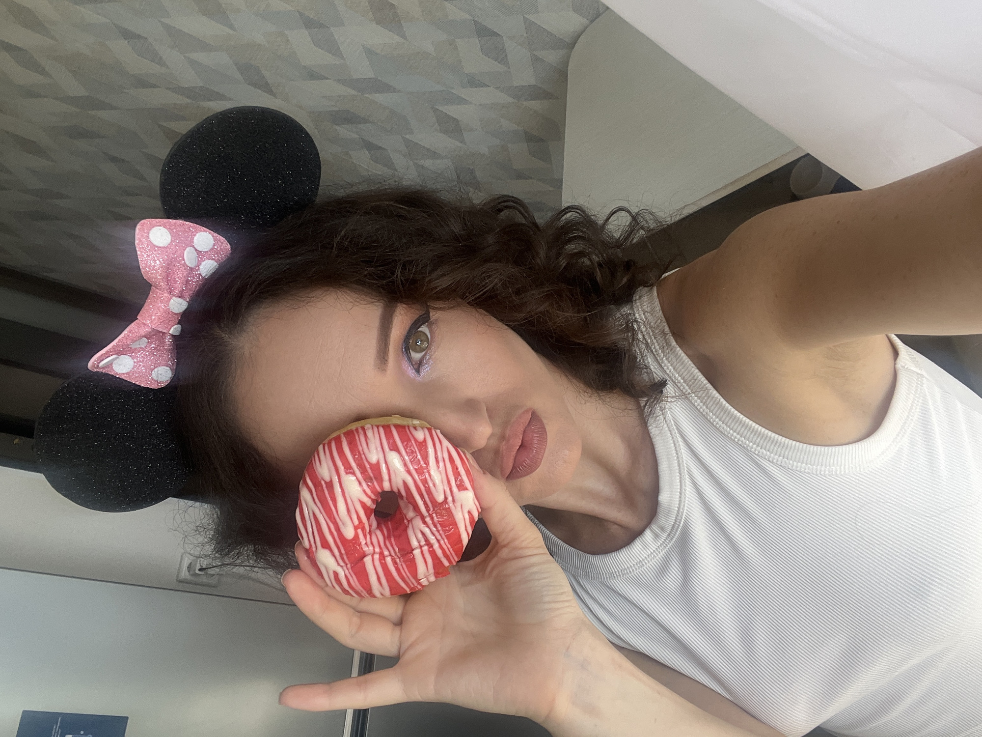 A woman with curly brown hair and wearing a white ribbed tank top and Minnie Mouse ears with a pink polka-dot bow is holding a donut in front of her face. The donut has red icing with white drizzles. She is making a kissing face. The background appears to be an indoor setting with a textured wall and a white surface. The image was taken in Kazan, Russia.