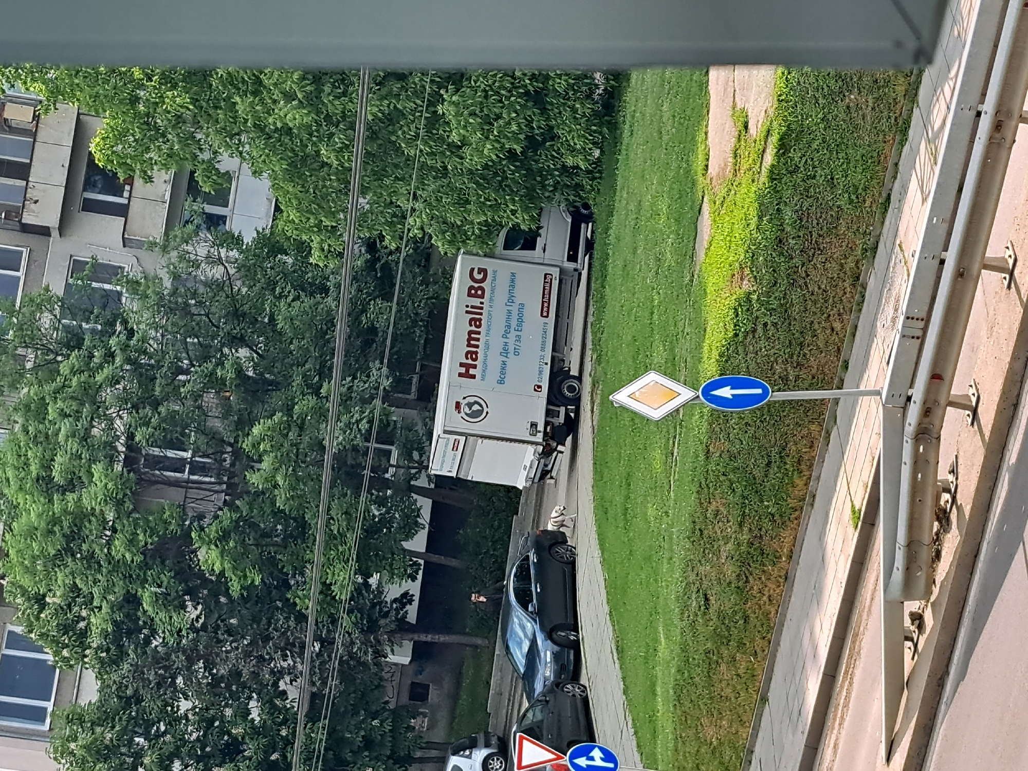 The image shows a view from a window or high vantage point, capturing a street scene. A building is visible to the left, partially obscured by lush green trees. A delivery truck with the branding "Hamali.BG" parked on the side of the road near a grassy area. Several parked cars are visible. The street is marked with a road sign indicating directions. The overall scene appears to be in an urban residential area.