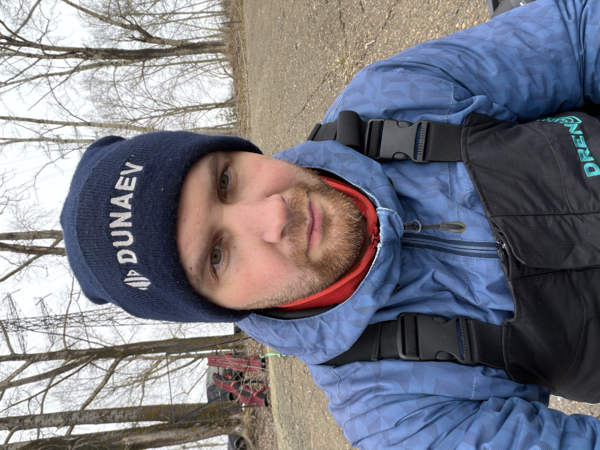 A man is taking a selfie outdoors. He is wearing a blue beanie with white lettering and a logo that reads "DUNAEV," a blue patterned jacket with a red undershirt visible at the neck, and black bib overalls with the word "DRENNAN" vertically printed on the right side. Black buckles are visible on the overalls. The man has light skin, a short beard, and light-colored eyes.
The background shows leafless trees and what appears to be a gravel or dirt path. A glimpse of a red and black object, possibly a piece of equipment or vehicle, is visible behind the man and to the left. The overall lighting suggests it is an overcast day.
Based on the clothing and the style of the surrounding environment, the location is likely a rural or semi-rural area in Chaikovsky, Russia, as the DUNAEV brand is associated with fishing equipment.