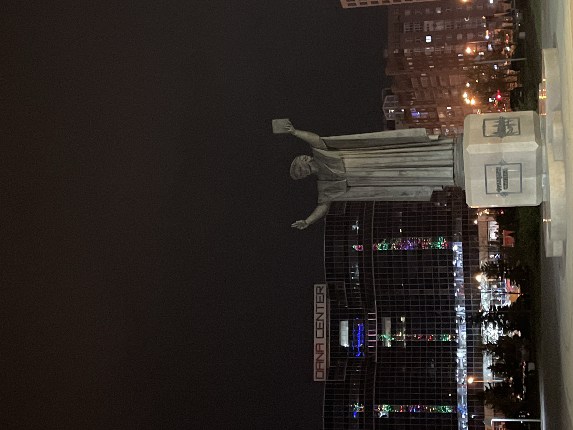 The media file displays a nighttime urban scene, featuring a prominent statue against a backdrop of illuminated buildings.
The central subject is a large statue of a robed male figure, possibly a historical or religious personality, standing on a hexagonal pedestal. The figure holds an object resembling a book in its right hand while its left arm is extended downwards. The statue is well-lit, appearing light-colored, possibly stone or marble, contrasting with the dark night sky.
In the background, several multi-story modern buildings are visible, their windows glowing with interior lights and reflecting colorful light patterns, suggesting either architectural lighting or nearby festive decorations. A distinct sign on one of these buildings clearly reads "DANA CENTER". Scattered streetlights and other urban illumination contribute to the nighttime ambiance. Small patches of greenery, likely trees or bushes, can be seen near the base of the buildings.
On the statue's hexagonal pedestal, two emblems or inscriptions are visible. The upper one appears to be a silhouette of a person on horseback. The lower emblem is a crest containing Cyrillic text, which is largely unreadable but seems to begin with "ПРЫ".
The scene depicts a peaceful, well-lit public space or city square at night, highlighting the monument within a modern urban environment. There are no people visible, and no specific event is taking place beyond the display of the statue and the nocturnal city lights.