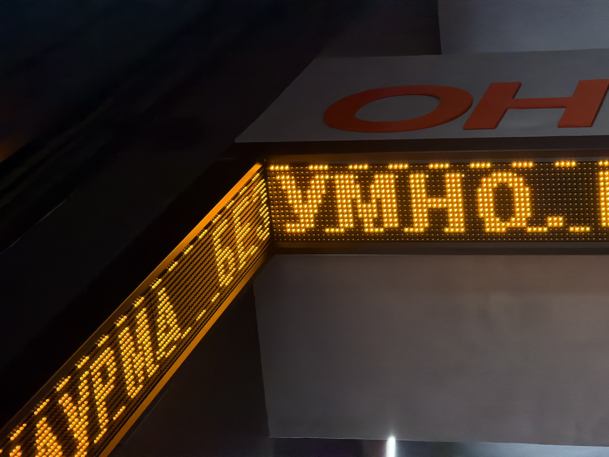 Captured at night, the image reveals an electronic display in Krasnoyarsk, Russia. The yellow LED sign reads "УМНОЖЕНИЕ" (multiplication), while a secondary sign to the left displays Cyrillic characters that are difficult to make out due to the perspective and angle of the shot. Above the display, partial orange letters are visible, possibly signage for the location. The scene's lighting comes primarily from the electronic sign, with the surrounding environment appearing dark.
