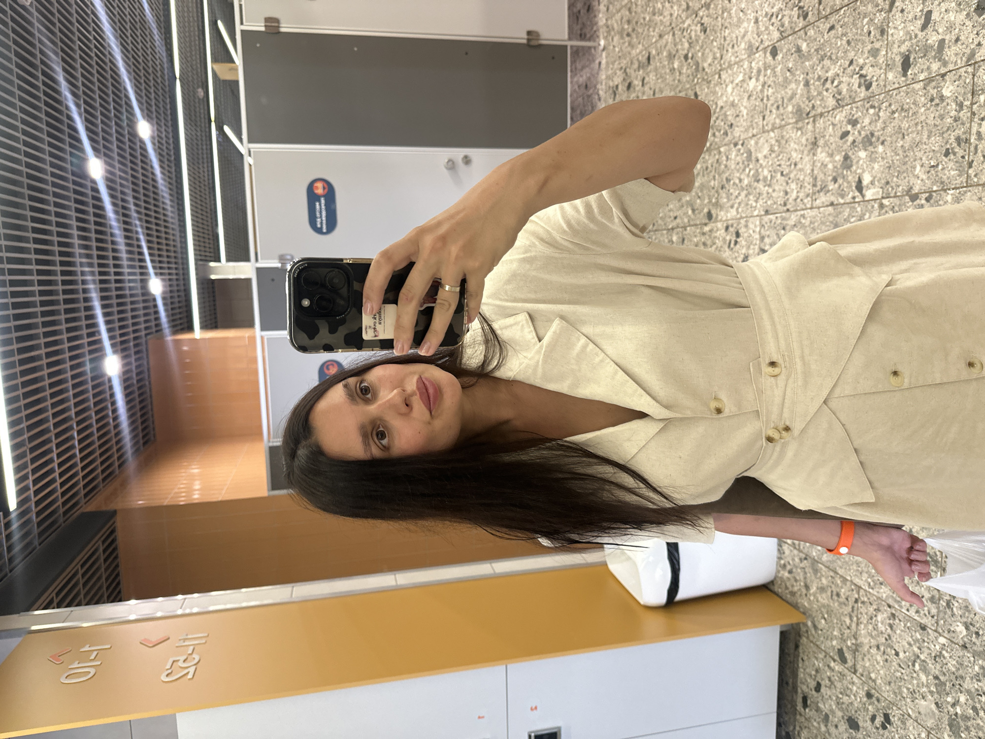A woman is taking a selfie in a modern interior space, possibly a public restroom or changing area, given the presence of stalls in the background. She is wearing a light-colored, collared dress with buttons and a cinched waist. She holds a smartphone in her right hand, which is angled to capture her reflection. On her left wrist, she wears a bright orange band. Her hair is long and dark, and she is looking towards the phone screen. The floor is tiled with a speckled pattern. To the left of the frame, there's a bright yellow vertical panel with some text in an unknown language, and a white trash bin. The lighting appears to be overhead, with visible ceiling grids and fixtures. The environment suggests a clean, functional public space. There is no indication of Votkinsk, Russia in the image itself.