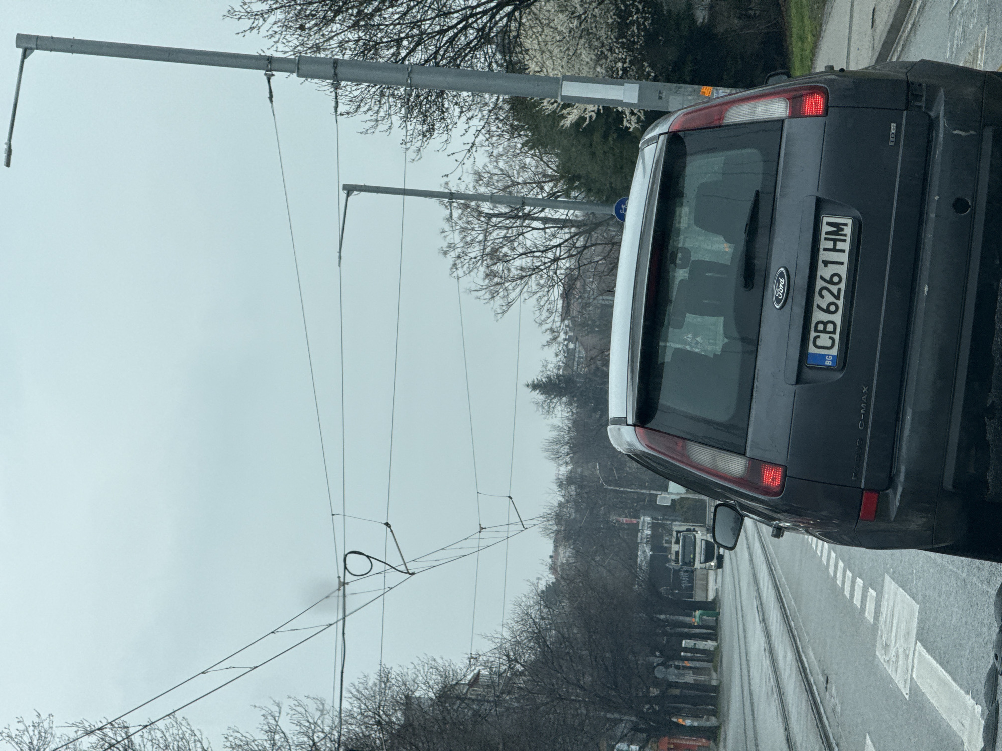 The image depicts the back of a dark gray Ford C-MAX car with a Bulgarian license plate "CB 6261 HM". The car is positioned on a road with tram tracks, and a crosswalk is visible. Overhead tram lines and supporting poles are prominent. Trees line the road. The weather appears overcast. Based on the vehicle's license plate code "CB", the location is Sofia, Bulgaria.
