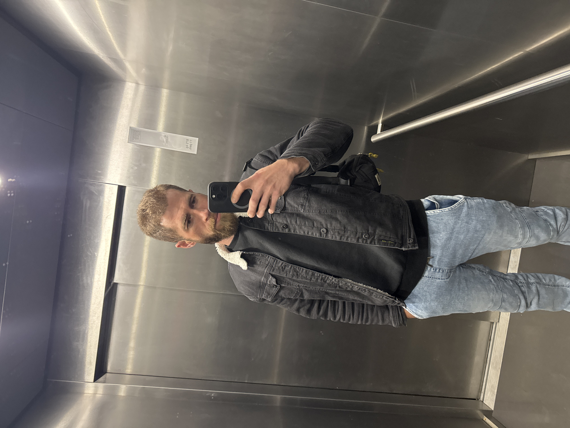 An adult male is observed inside an elevator, taking a selfie using a black smartphone. The elevator interior features brushed stainless steel walls and a dark, non-reflective floor. A horizontal cylindrical metal handrail is present on the right wall. A light grey, rectangular sticker or label is affixed to the right wall, displaying mirrored and inverted text, partially decipherable as "d4 258" and "2199 1f," likely indicating elevator identification. The male has light brown hair and a full beard. He is dressed in a black long-sleeved shirt, a dark grey or black denim jacket with a white/cream sherpa collar, and light blue washed denim jeans. His left hand is in his front left pocket, while his right hand holds the smartphone, which has a visible magnetic ring accessory on its back. A small black bag with yellow accents is slung over his left shoulder, resting on his back. The specific location within Budva, Montenegro, cannot be determined from the image alone.