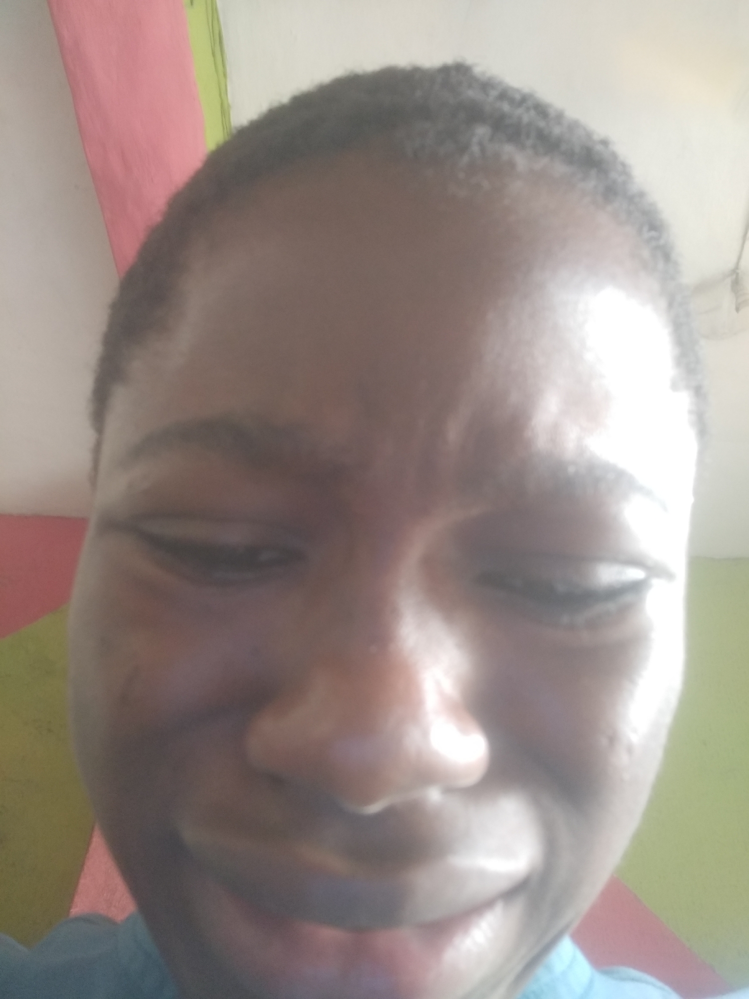 A close-up selfie of a dark-skinned individual with short, dark hair. The person is looking down with a slight frown, and the image is slightly blurry. The background shows portions of a wall painted in pink and green. The location is Shangisha, Nigeria.