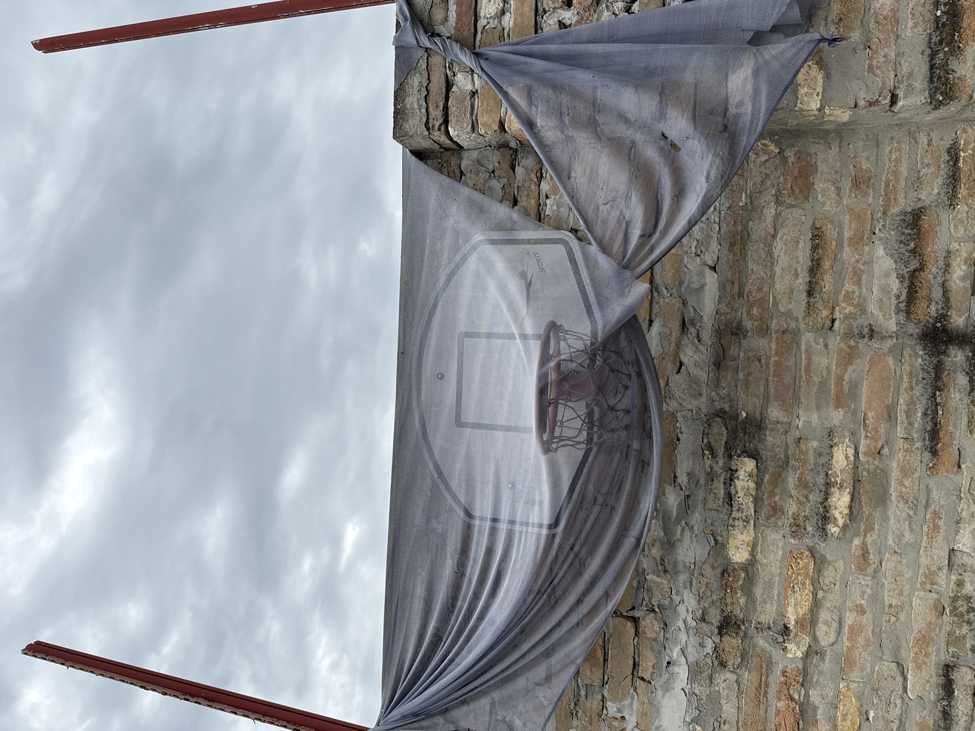 The image depicts a weathered basketball hoop and backboard mounted on a brick wall. A grey, tattered net or tarp is draped over the backboard and extends down the side, partially obscuring the view. Two red metal posts are visible on the left and right edges of the frame, suggesting a structure associated with the basketball setup. The brick wall itself shows signs of age and wear, with some crumbling mortar and discolored bricks. The background consists of a cloudy, overcast sky. The scene appears to be located in an outdoor urban or semi-urban environment, possibly a neglected recreational area or a yard within Apatin Municipality, Serbia. There are no people or animals visible in the image. The overall impression is one of disuse and decay.