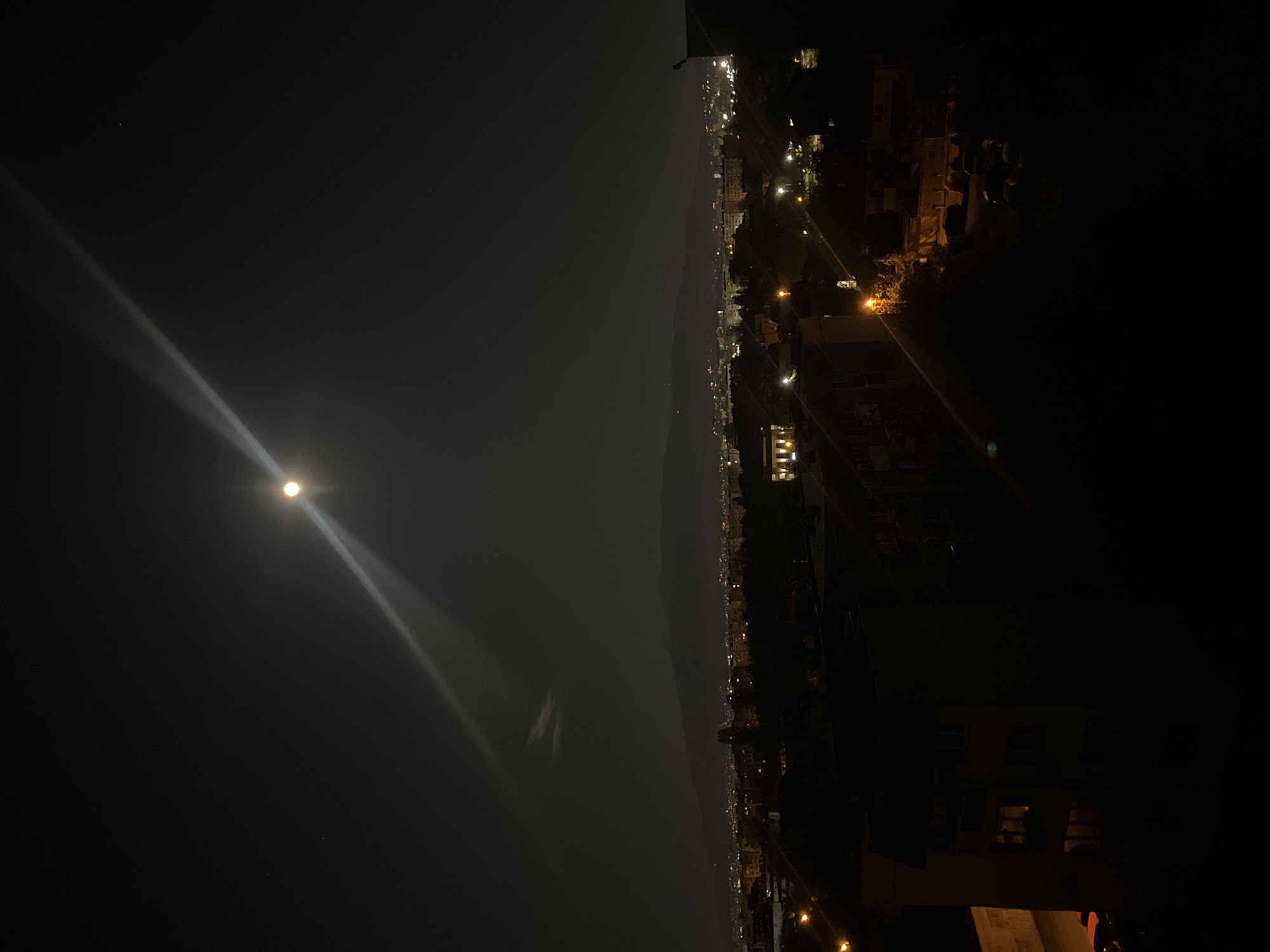 The image depicts a night scene overlooking what appears to be the city of Sofia, Bulgaria. A bright moon dominates the sky on the left, casting a strong beam of light. Below the moon, a body of water or large open space can be seen. In the center, the silhouette of distant mountains separates the open space from the urban landscape of Sofia, where numerous city lights are visible. On the right, the foreground is dominated by the dark shapes of buildings, possibly apartments or commercial structures. The overall atmosphere is dark and illuminated only by the moon and the artificial lights of the city. No people are visible in the image.