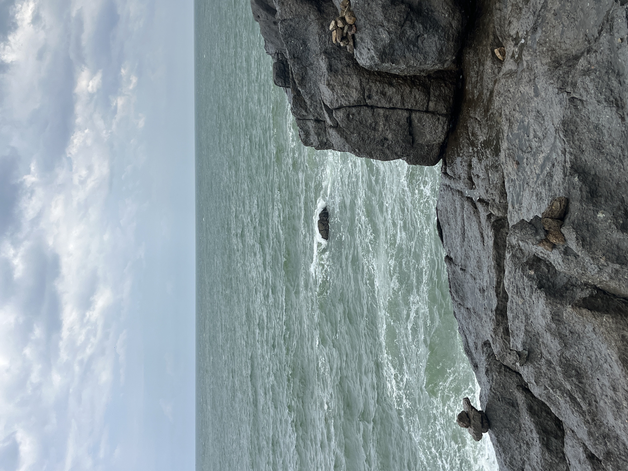 The image depicts a rocky coastline on an overcast day. The sea is a muted green, with whitecaps indicating a moderate chop. In the foreground, dark, textured rocks form the shore, with pools of water reflecting the sky. Several cairns, stacks of balanced rocks, are visible on the rocks. One cairn, near the left edge of the image, is composed of three rocks. Another, on the right side of the image, is a larger stack of about seven rocks. The horizon line is clear, with a band of grey clouds dominating the upper portion of the frame. The scene is devoid of any people or man-made structures other than the cairns. This location is the Red Sand Dunes area in Mui Ne, Vietnam, known for its coastal cliffs and unique geological formations.