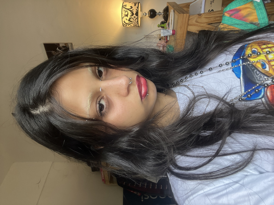 The image is a medium close-up portrait of a young individual, presented rotated 90 degrees counter-clockwise. The subject's head is positioned to the left, and their body extends to the right.
The person has long, dark, wavy hair that is parted slightly off-center and cascades over their shoulders and chest. Their skin is fair with visible freckles across the bridge of their nose and cheeks. Their eyebrows are bleached or very light in color, providing a strong contrast against their dark hair. They have dark eyes that appear to be looking towards or slightly past the camera. Their lips are adorned with glossy red lipstick.
Several facial piercings are prominent:
* A silver horizontal bar piercing (bridge piercing) is located between their eyes at the top of their nose.
* A silver stud (nostril piercing) is on the left side of their nose.
* A silver ring (septum piercing) passes through the septum of their nose.
* A silver stud (philtrum or Medusa piercing) is centered above their upper lip.
* Two silver studs (spider bite piercings) are positioned on the left side of their lower lip.
* Another silver stud is visible directly below the center of their lower lip, possibly a vertical labret.
The person is wearing a white t-shirt with a colorful graphic design visible on the right side. The design depicts what appears to be an animated character's head or a face, featuring colors such as yellow, blue, and red. Multiple strands of silver-colored beaded necklaces, varying in length and bead size, are draped over the white t-shirt, with one resembling a rosary-style chain.
The background suggests an indoor setting, likely a personal room. In the upper background, a wall-mounted light fixture with a black and white patterned lampshade casts a warm, yellowish glow. Below this, a wooden structure, possibly a shelf or headboard, holds various items including a decorative table lamp with a black base and silver elements, some stacked books or boxes, and a green and multi-colored fabric item. A dark-colored picture frame or poster is partially visible on the wall in the upper left background.
The overall impression is one of individual style and personal expression.
Visible text includes a partial view of "Pepsi" in white letters against a dark background in the lower right corner and "LONDA" on a label or box in the lower left corner (as seen in the cropped hints).