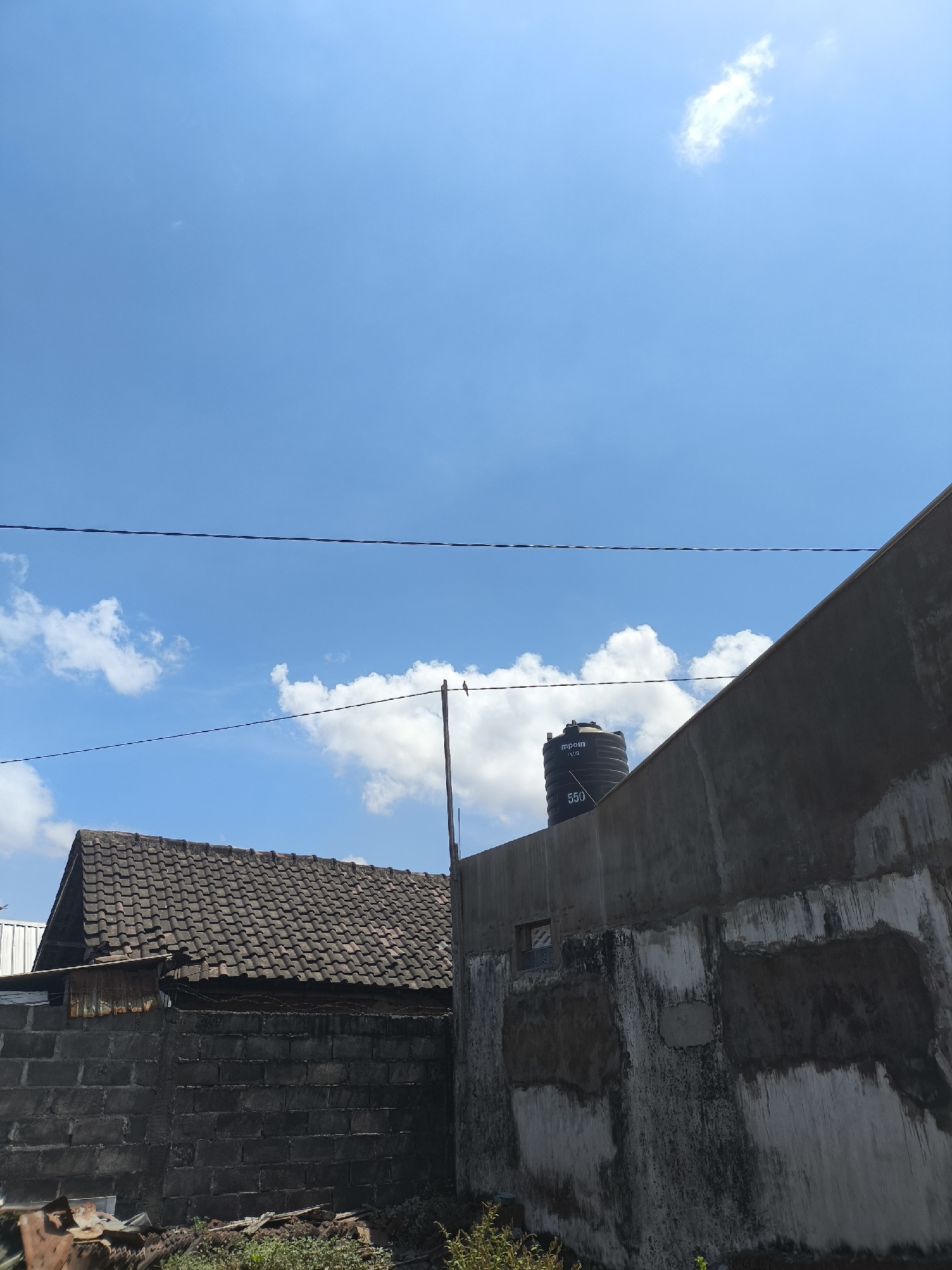 The image shows a partly built or dilapidated structure in an outdoor setting, likely in Kebonsari, Indonesia. The sky is a vibrant blue with scattered white clouds. In the foreground, a dark brick wall is visible, with some debris and vegetation in front of it. To the left, a house with a tiled roof appears to be older and less maintained, with some corrugated metal sheeting attached to the side. A water tank, black and labeled "mpurn plus 550," is mounted on top of a concrete structure to the right of the house. This concrete structure has a rough, unfinished texture and appears to be part of a larger building. A thin wire runs horizontally across the frame, bisecting the sky and the structures below. Another similar wire is positioned lower, above the house. A small, dark object, possibly a bird, is perched on a thin pole to the right, near the water tank. The overall impression is one of an everyday, semi-urban or rural scene in Indonesia, with elements of construction, habitation, and the natural environment.