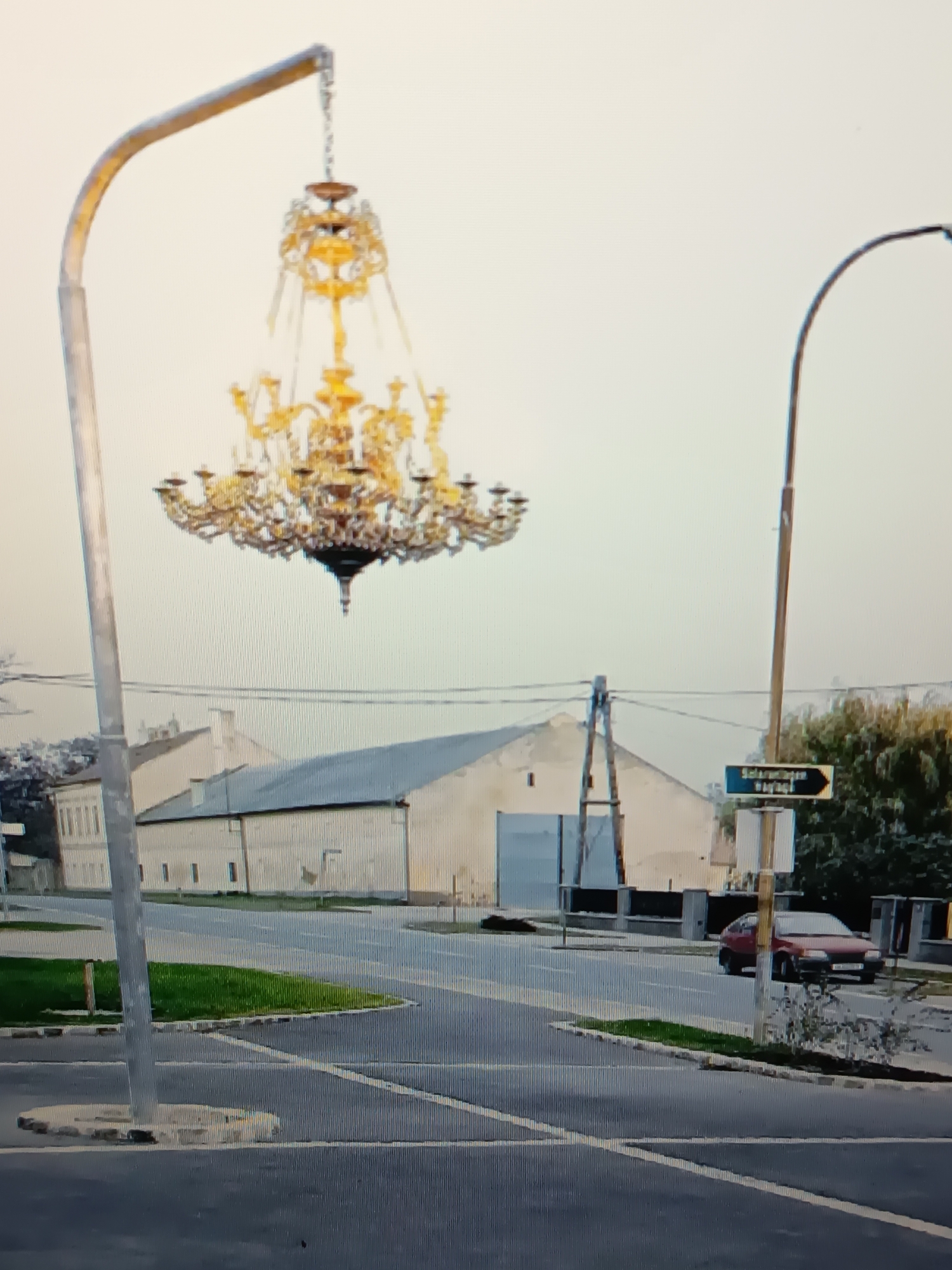 In the city of Serhiivka, Ukraine, an ornate, golden chandelier hangs suspended from a street lamp on a bright, overcast day. The street lamp, a slender, curved metallic pole, extends from the left side of the frame. The chandelier itself is elaborate, featuring multiple tiers of arms with candle-like fixtures and numerous dangling crystals, catching what little light is available in the sky.
Below the chandelier, a wide, paved intersection is visible. The road surface is dark asphalt, marked with white and yellow lines indicating lanes and pedestrian crossings. To the left of the intersection, a strip of vibrant green grass borders the roadway, delineated by concrete blocks. Further into the background, a long, low-slung building with a light-colored facade and a grey roof dominates the scene. This building appears to be industrial or utilitarian in nature, with large, possibly closed, doorways. A smaller, lighter-colored building with multiple windows is visible to the far left.
On the right side of the intersection, another street lamp, taller and straighter than the first, is positioned. Attached to this lamp is a blue street sign with white lettering and an arrow pointing to the right. The text on the sign is partially legible but appears to be in a Cyrillic alphabet. Beyond the sign, a cluster of green trees and foliage adds a natural element to the otherwise urban landscape. A red hatchback car is seen driving on the road, approaching the intersection from the right. The overall environment appears to be a suburban or semi-industrial area.