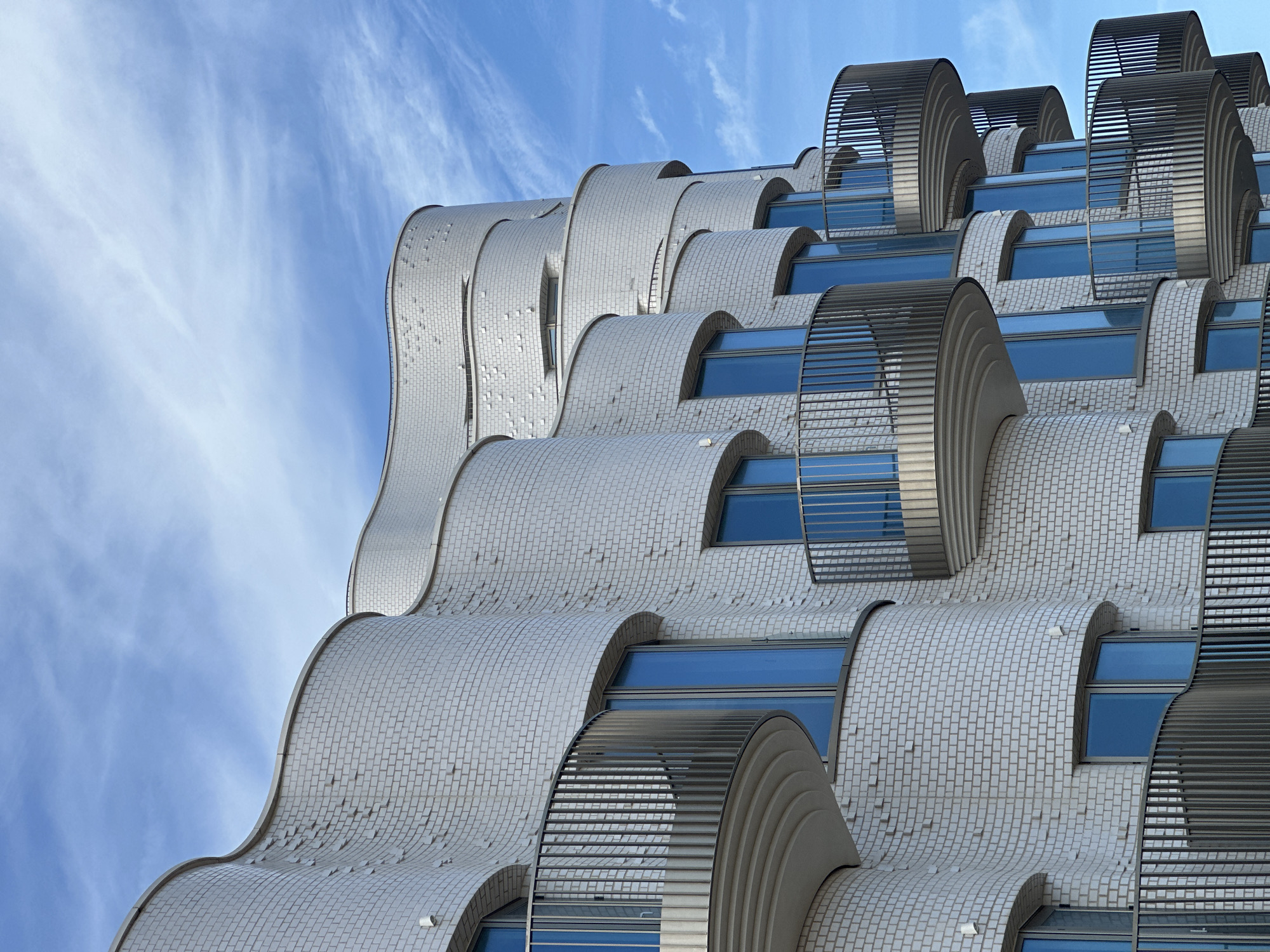 The image shows a close-up of the exterior of a modern building. The building is made of light grey bricks and features undulating, wave-like protrusions with windows. These protrusions are partially covered by curved, slatted metal screens. The background is a bright blue sky with light, wispy clouds. This building is located in the Folkestone and Hythe District, United Kingdom.